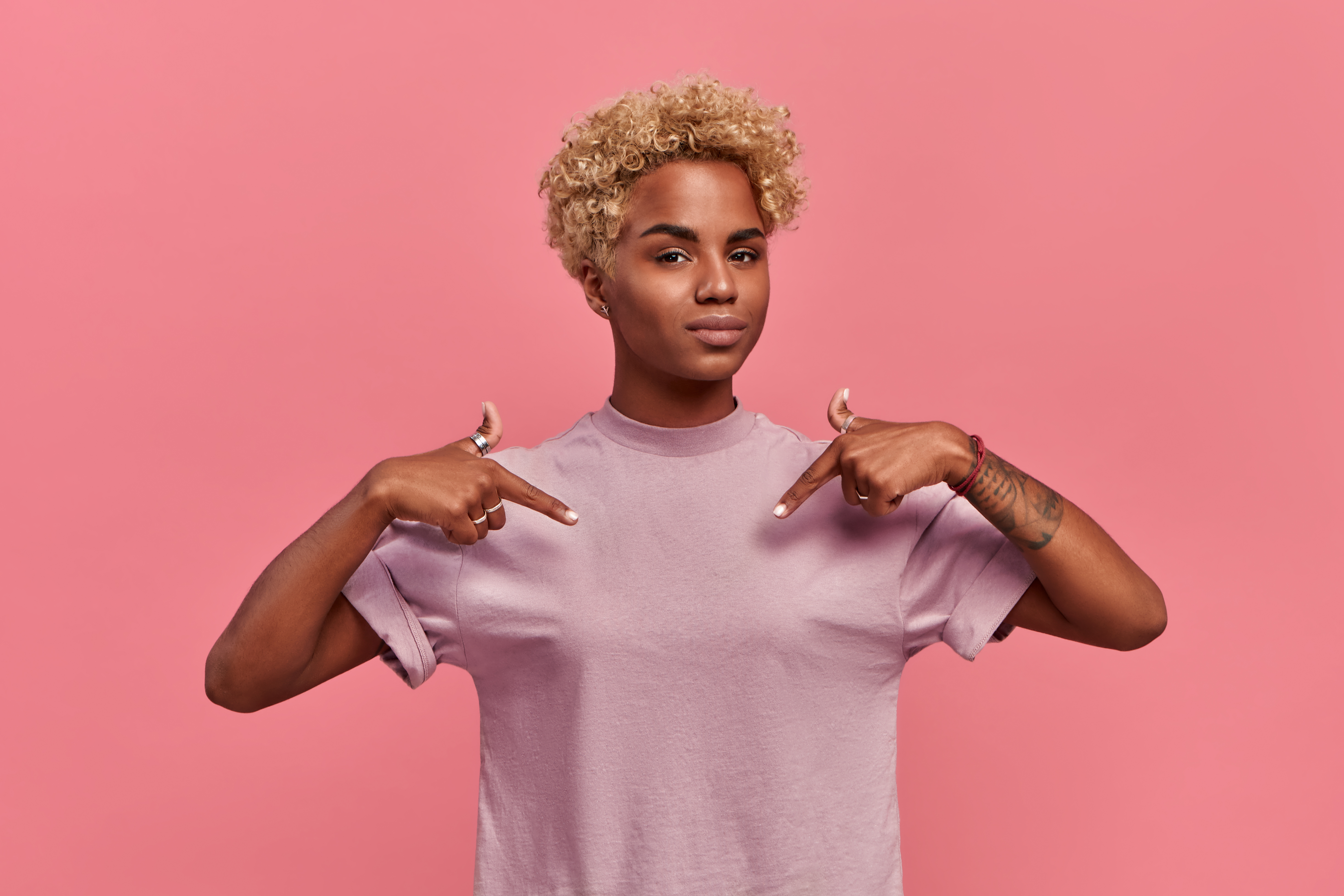 Selbstbewusste, stolze afroamerikanische Frau mit blonder Frisur, die mit ihren eigenen Erfolgen zufrieden ist, zeigt mit zwei Händen auf sich selbst oder auf ein T-Shirt zum Bedrucken, isoliert auf rosa Hintergrund | Quelle: Getty Images