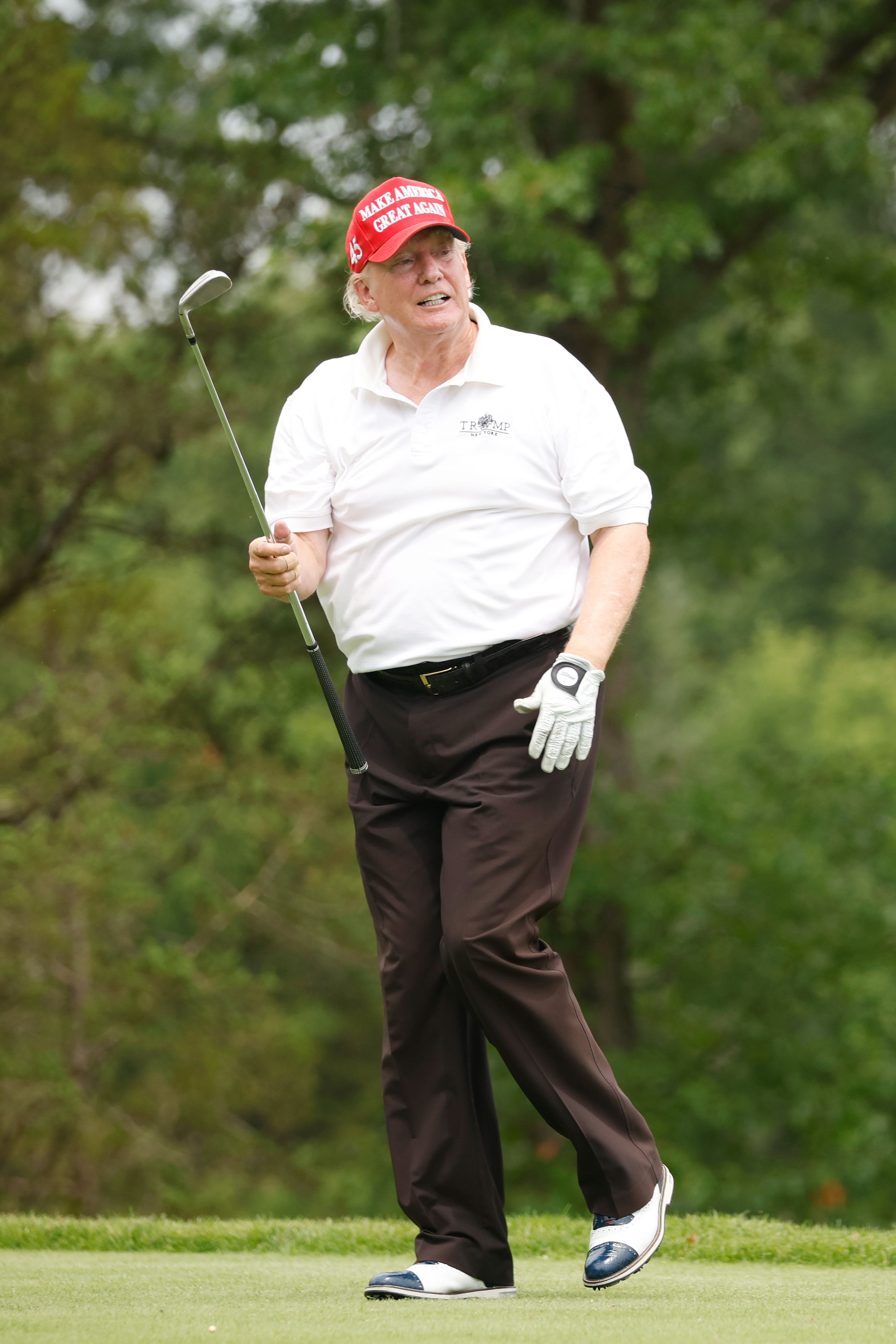 Donald Trump wirkt entspannt, mitten im Schritt, hält mit einer Hand einen Schläger und lächelt leicht. Die ungleichmäßigen Hauttöne sind deutlich zu erkennen, besonders um sein Kinn und seine Schläfen herum.