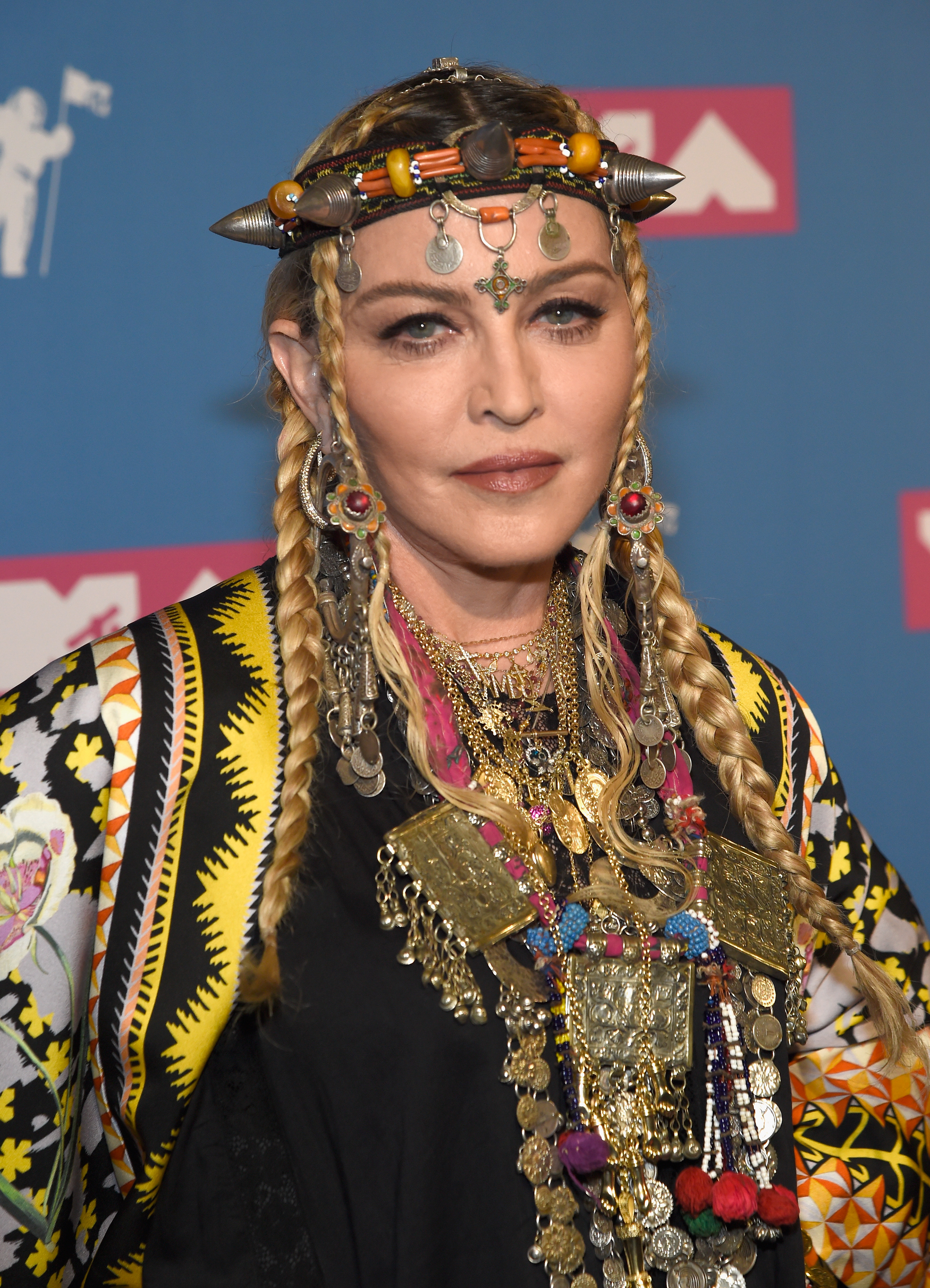 Madonna blickt mit langen blonden Zöpfen, einem kunstvollen Kopfschmuck, übergroßen Ohrringen, mehrreihigem Schmuck und einem Outfit mit bunten Mustern in die Kamera. | Quelle: Chat GPT