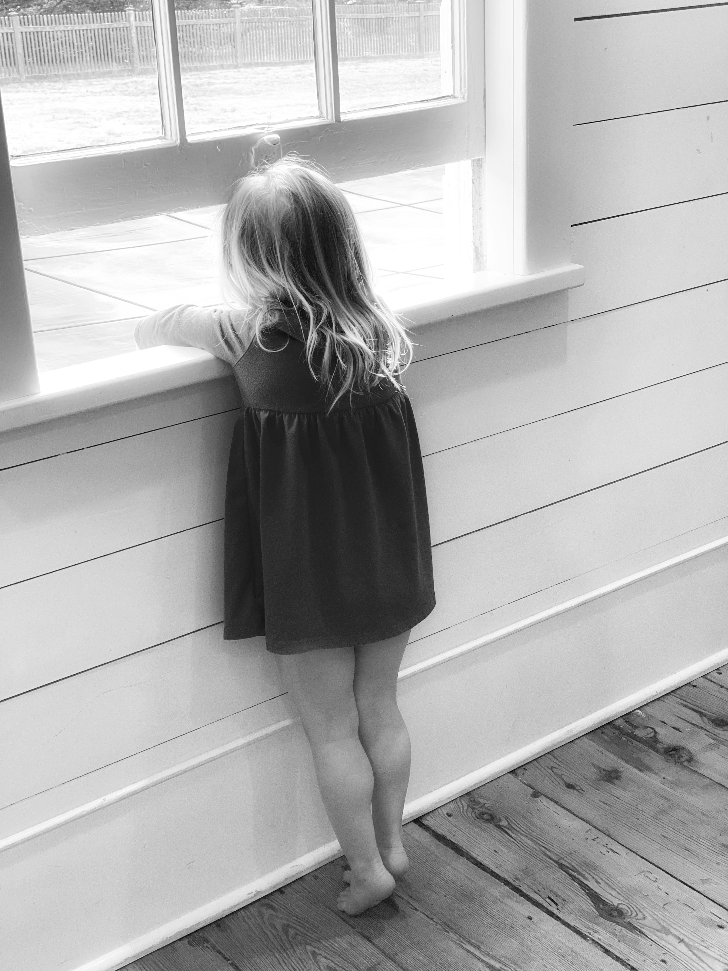 Eine Graustufenaufnahme eines kleinen Mädchens, das barfuß steht und aus dem Fenster schaut | Quelle: Unsplash