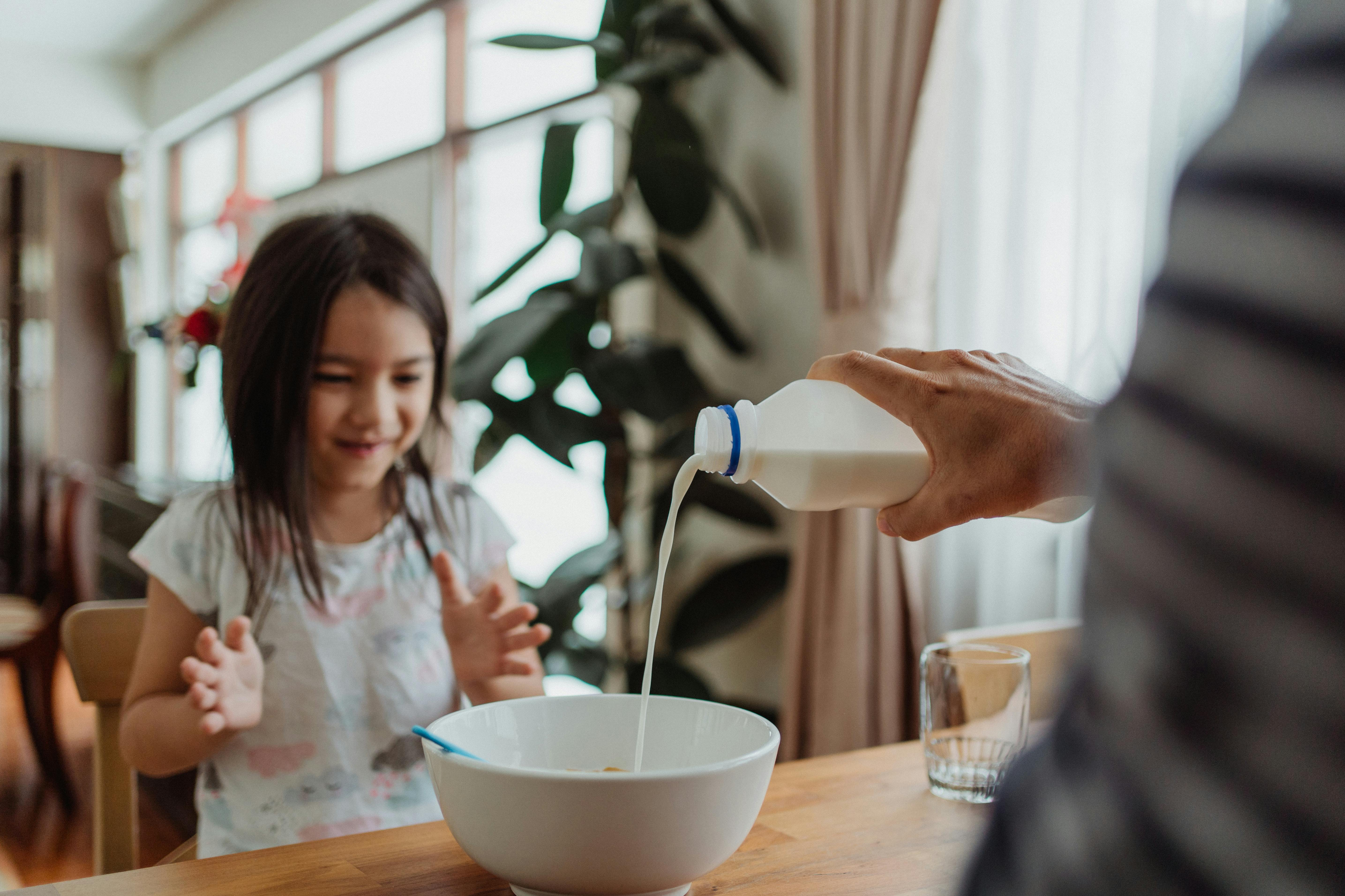 Ein Mann gießt Milch in eine Schüssel für ein junges Mädchen am Frühstückstisch | Quelle: Pexels