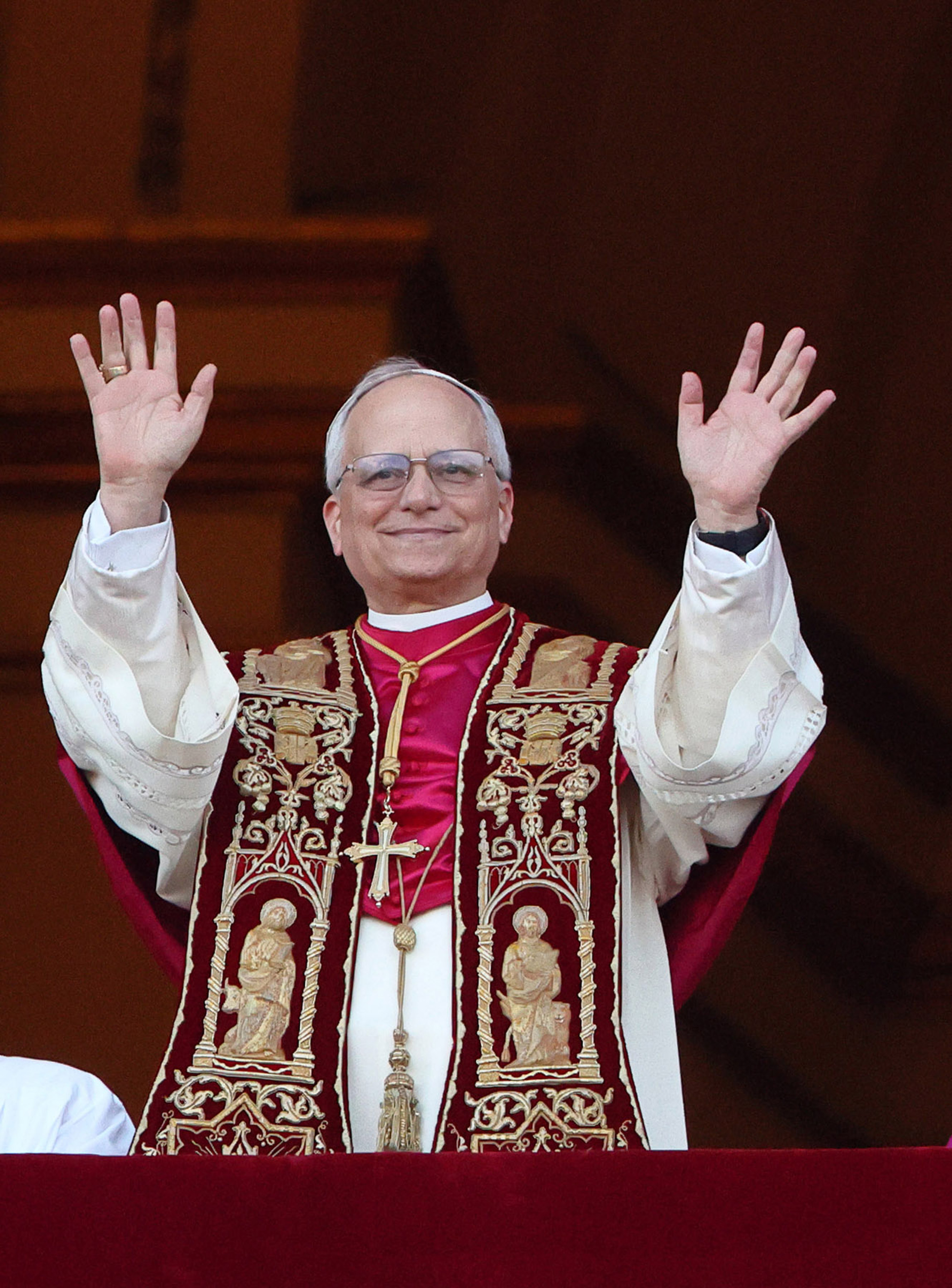 Papst Leo XIV. lächelt warm, als er beide Hände zur Begrüßung hebt. Seine verzierte karmesinrote Stola mit den goldenen Heiligen fängt das Licht ein