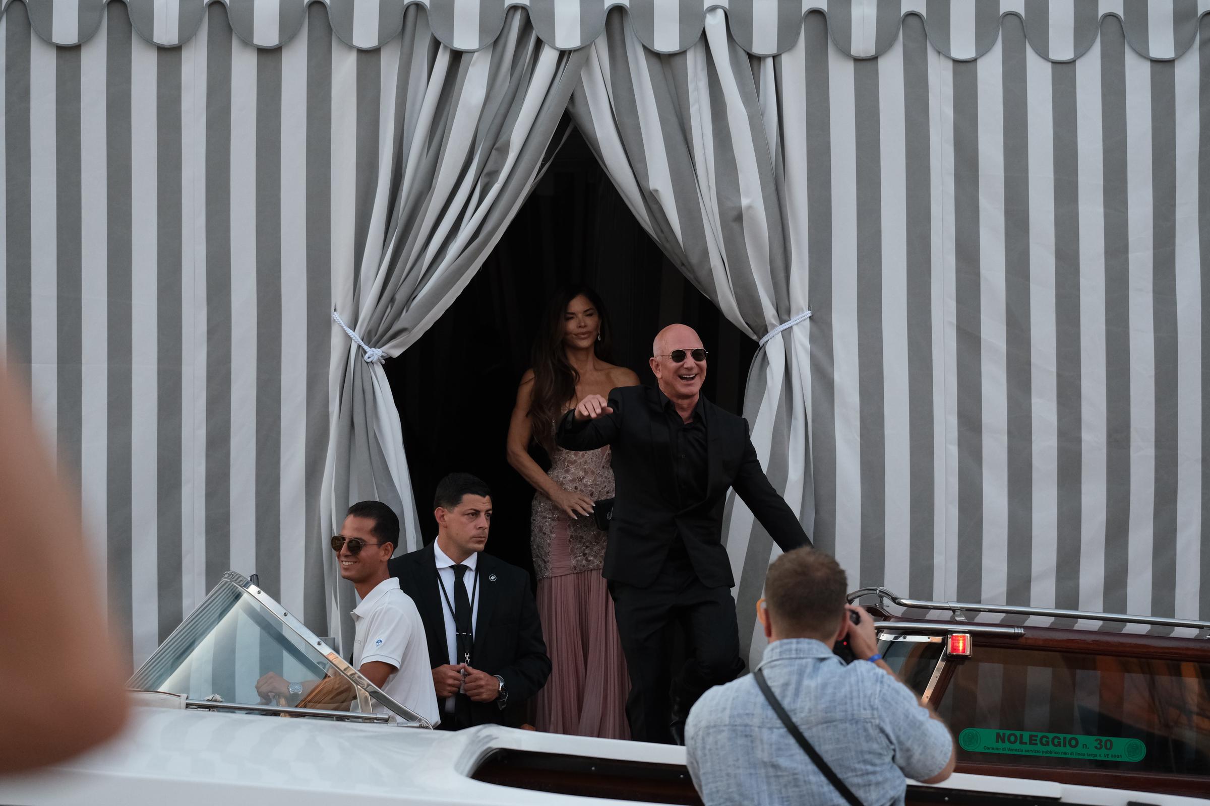 Jeff Bezos strahlt und winkt begeistert, während er in ein Wassertaxi steigt. Lauren Sánchez folgt ihm in einem errötenden Perlenkleid, eingerahmt von gestreiften Vorhängen und umgeben von Mitarbeitern und Schaulustigen.