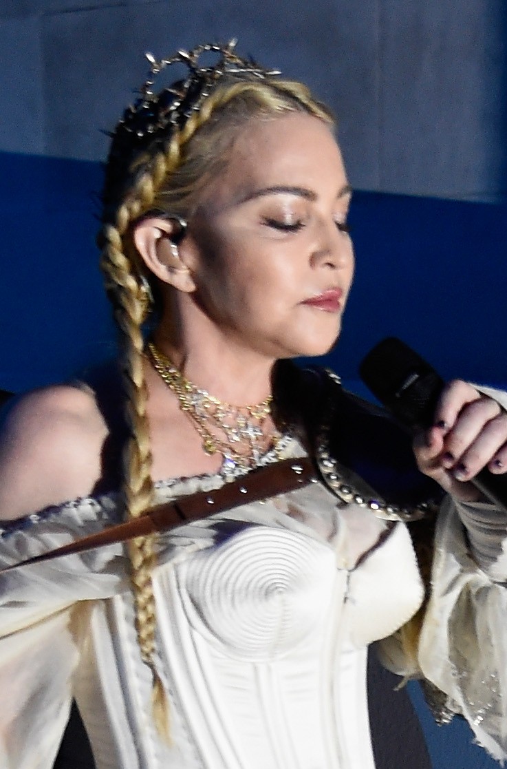 Madonna tritt mit langen blonden Zöpfen, mehreren Halsketten und einem weißen Korsettkostüm auf und hält ein Mikrofon, während sie nach unten blickt. | Quelle: Chat GPT