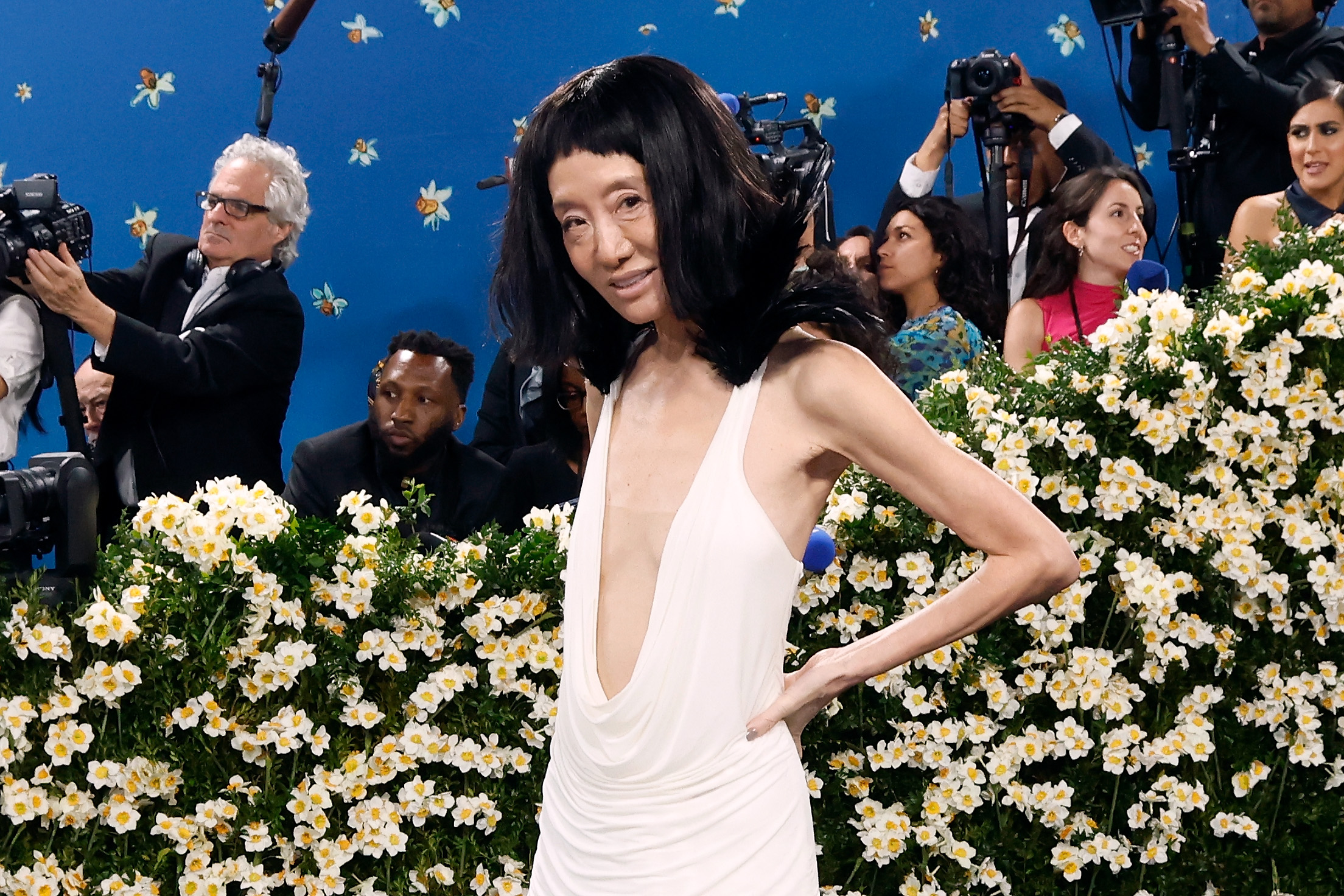 In selbstbewusster Pose steht Vera Wang vor einem Feld aus weißen Blüten, die den tiefen Ausschnitt und die gefiederten Schultern ihres maßgeschneiderten Kleides betonen.
