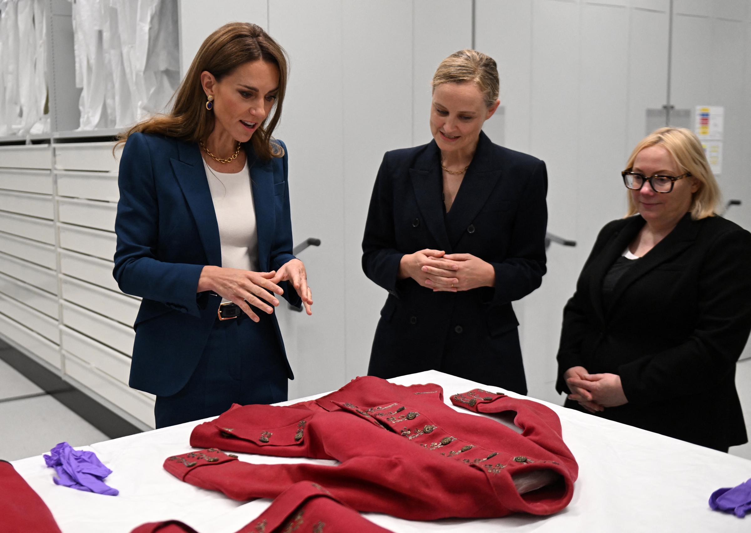 Prinzessin Catherine begutachtet zusammen mit den Kuratorinnen Oriole Cullen und Kate Parsons ein historisches rotes Reitkleid. Konzentriert und engagiert gestikuliert sie nachdenklich, während sie über die feinen Details des Kleidungsstücks und die Erhaltung der Textilien spricht.
