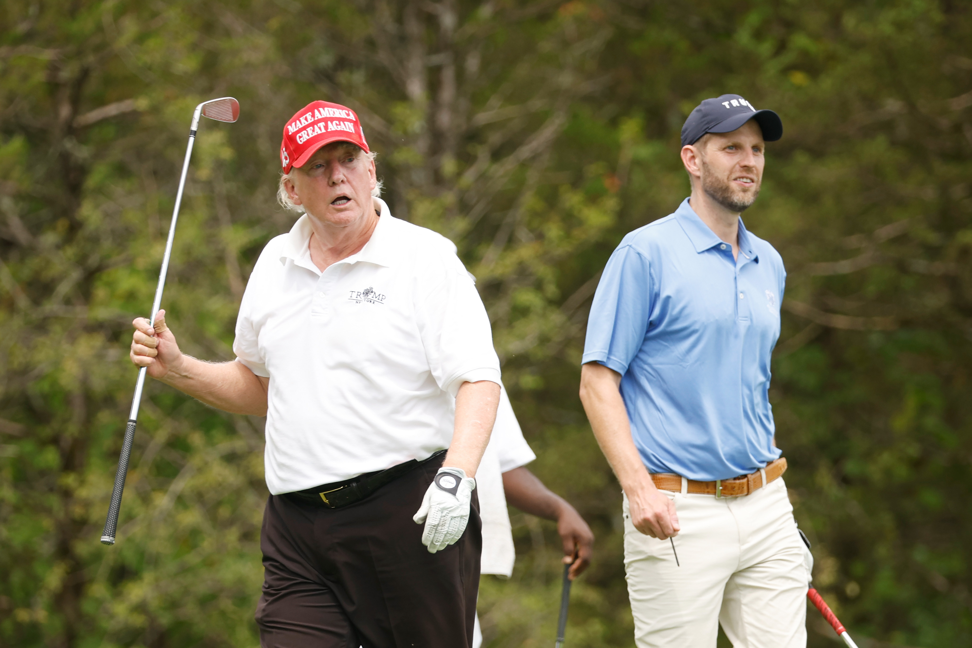 Donald Trump läuft während des Turniers an der Seite seines Sohnes Eric Trump und hält dabei einen Schläger in der Luft. Sein Gesicht zeigt sichtbare Rötungen an den Wangen und der Nase, während der Rest seiner Haut heller aussieht.