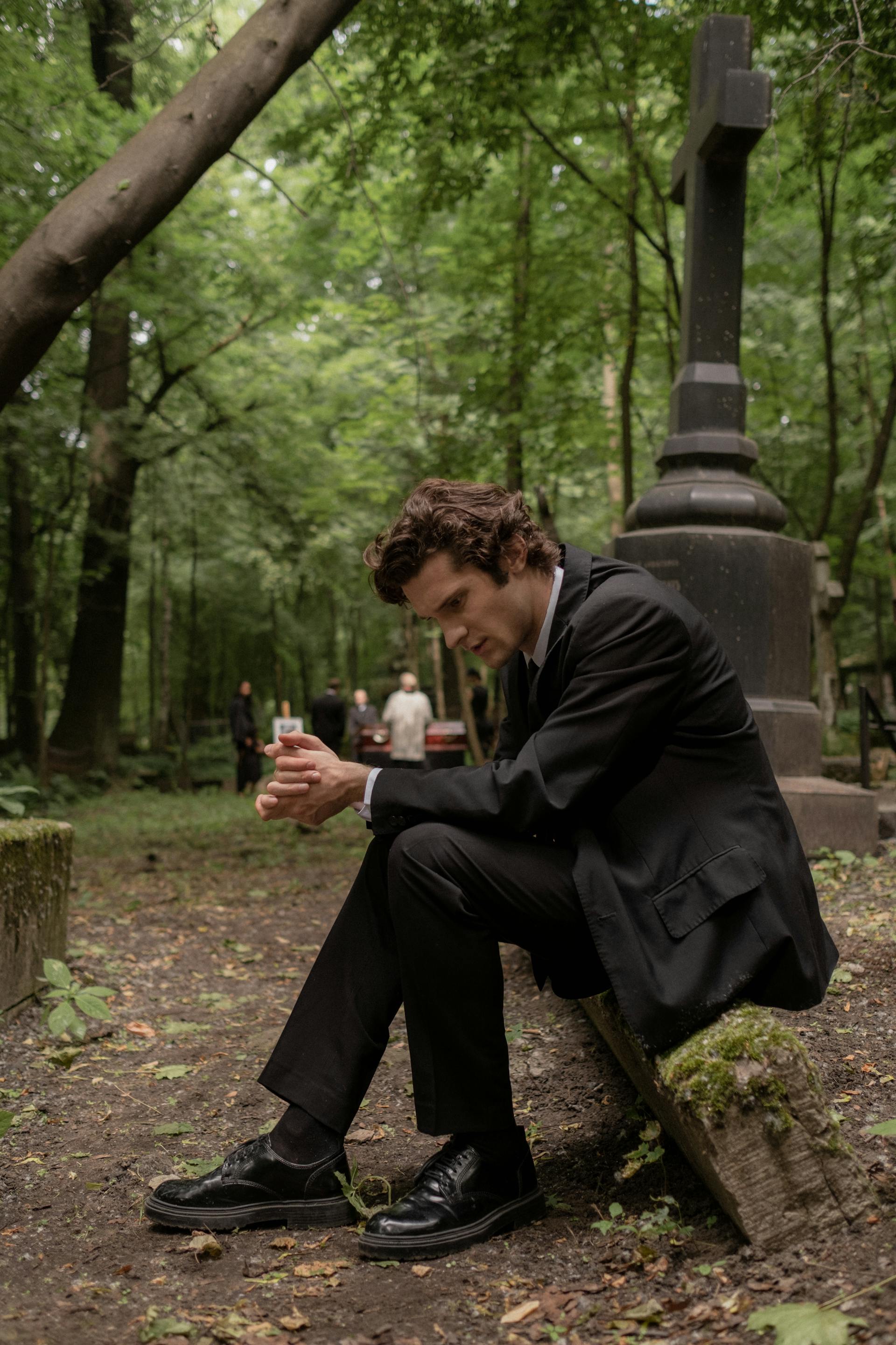 Ein Mann in schwarzem Anzug und Jacke sitzt auf einem Friedhof | Quelle: Pexels