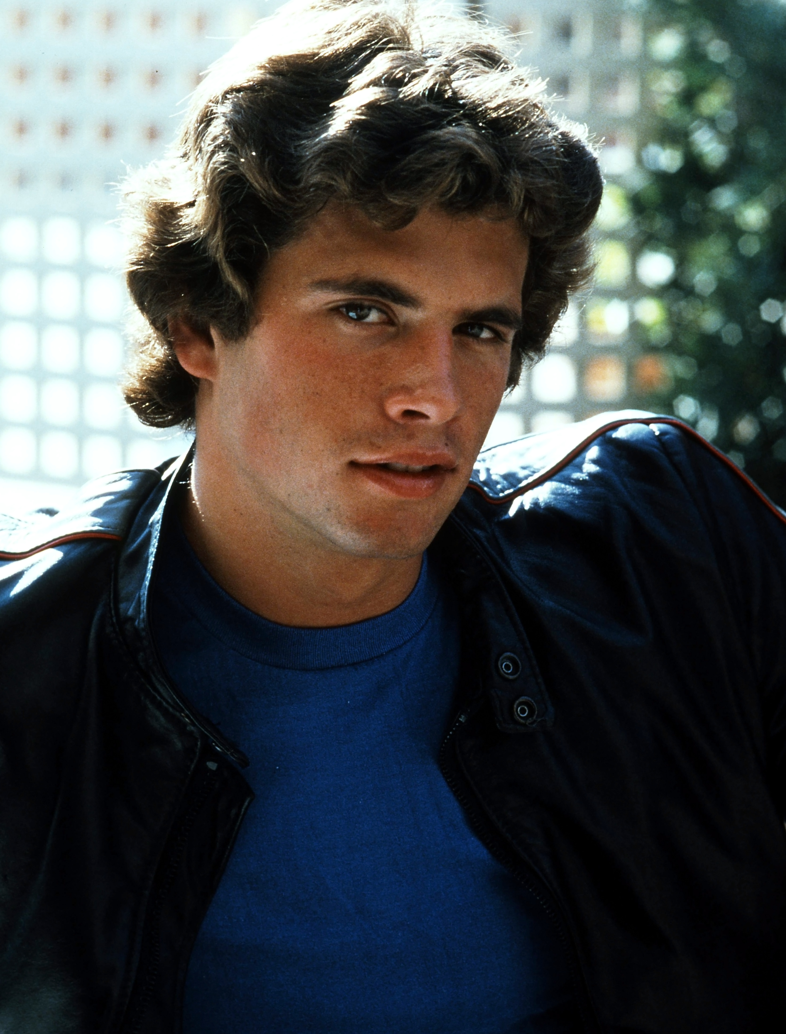 Der aufstrebende Schauspieler strahlt in einem farbigen Vintage-Porträt aus dem Jahr 1978 mühelose Coolness aus. Er trägt ein leuchtend blaues T-Shirt mit Rundhalsausschnitt unter einer schwarzen Lederjacke mit roten Ziernähten und blickt mit voluminösen, sonnengeküssten braunen Locken und markanten, kantigen Gesichtszügen in die Kamera. | Quelle: Getty Images
