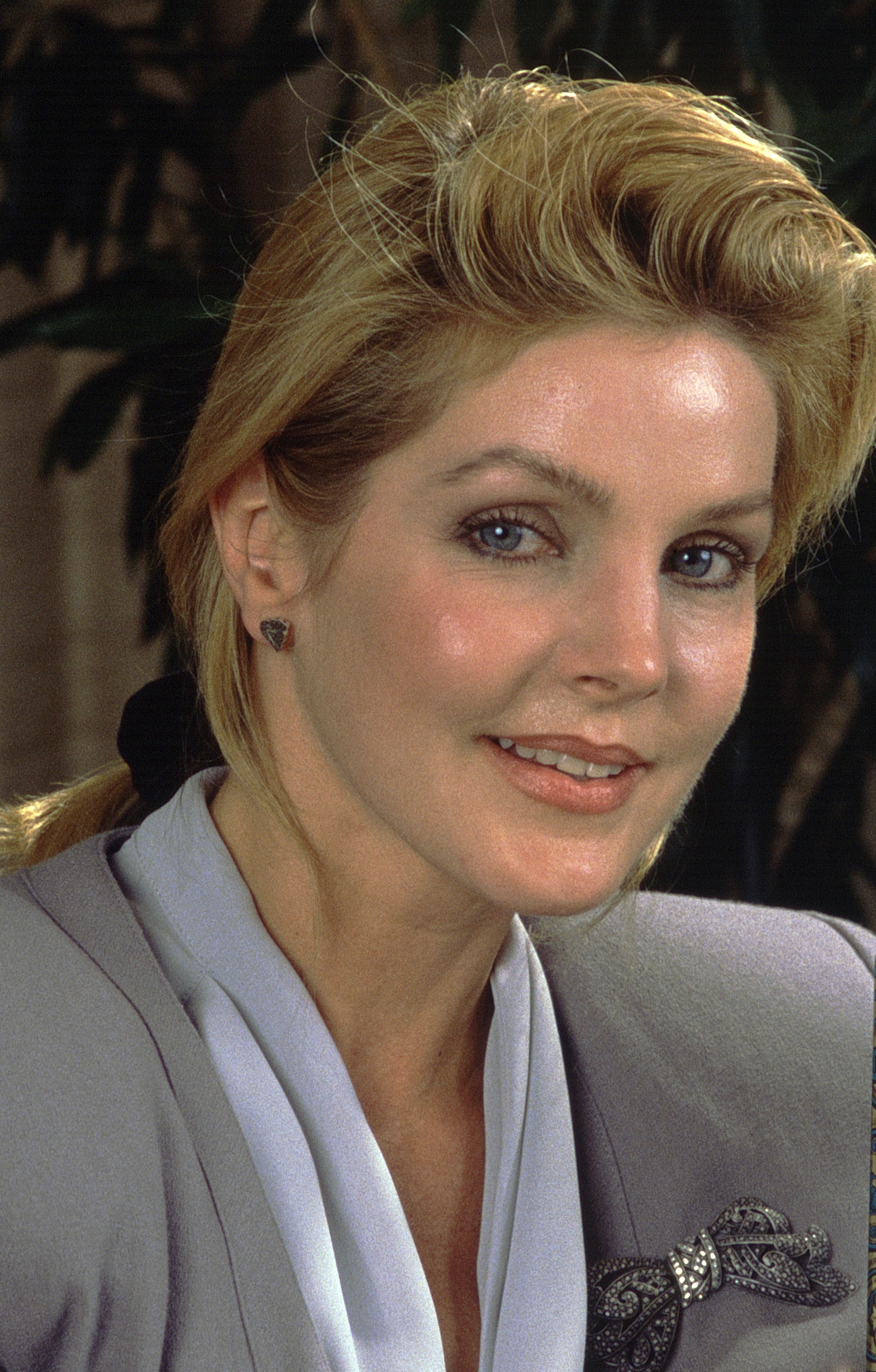 Priscilla Presley posiert für ein Porträt während der Vorstellung eines neuen Parfüms in Los Angeles, Kalifornien, am 13. Mai 1988. Sie trägt einen maßgeschneiderten grauen Blazer, der durch eine kunstvoll verzierte, mit Edelsteinen besetzte Schleifenbrosche akzentuiert wird, darüber eine weiche, weiß drapierte Bluse. Ihr blond gesträhntes Haar ist zu einem tief sitzenden Pferdeschwanz zurückgebunden, der mit einem dunklen Haargummi befestigt ist. | Quelle: Getty Images