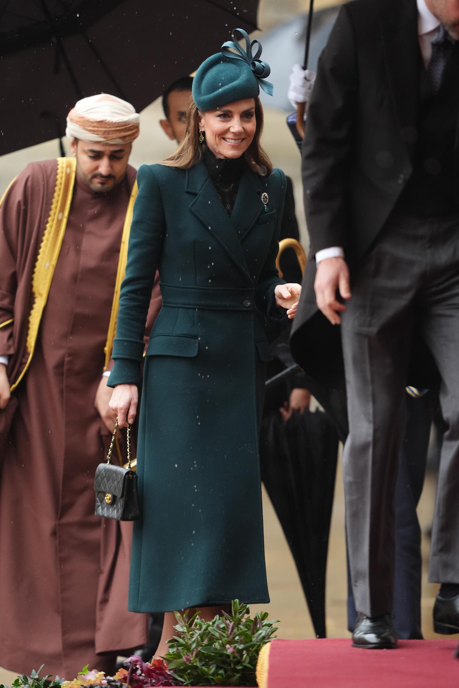 Catherine, Prinzessin von Wales, lächelt sanft, während sie am 20. Oktober 2026 über das feuchte Festgelände in Windsor schreitet. Sie trägt ein strukturiertes, waldgrünes Mantelkleid, das durch ein zartes, schwarz-spitzenbesetztes Unterkleid mit hohem Kragen und die historische „Queen Alexandra Three Feathers“-Brosche am Revers akzentuiert wird, und hält eine kleine, schwarze, gesteppte Chanel-Klappentasche in der Hand, während sie neben Prinz William geht. | Quelle: Getty Images
