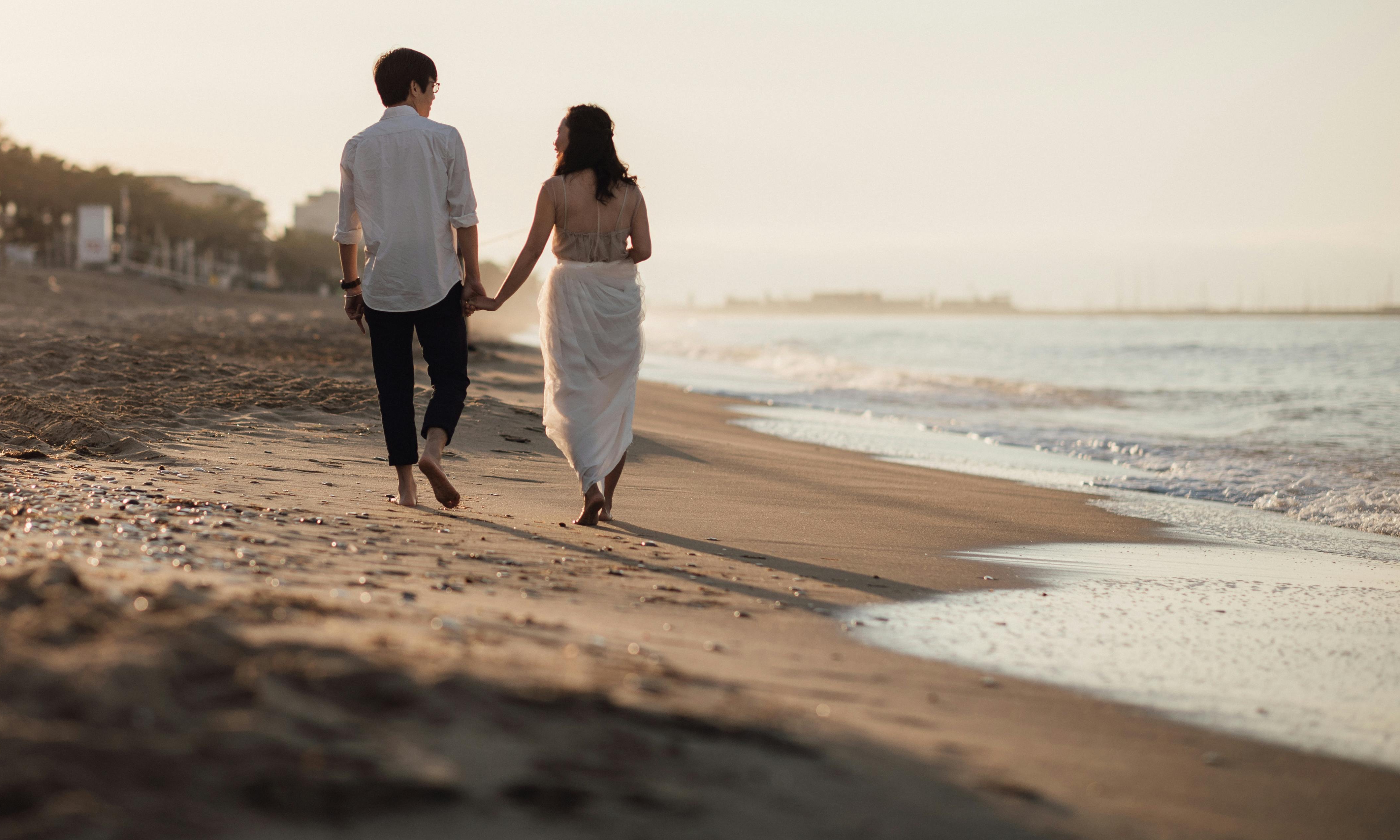 Ein Paar, das bei Sonnenuntergang Hand in Hand am Strand spazieren geht | Quelle: Pexels