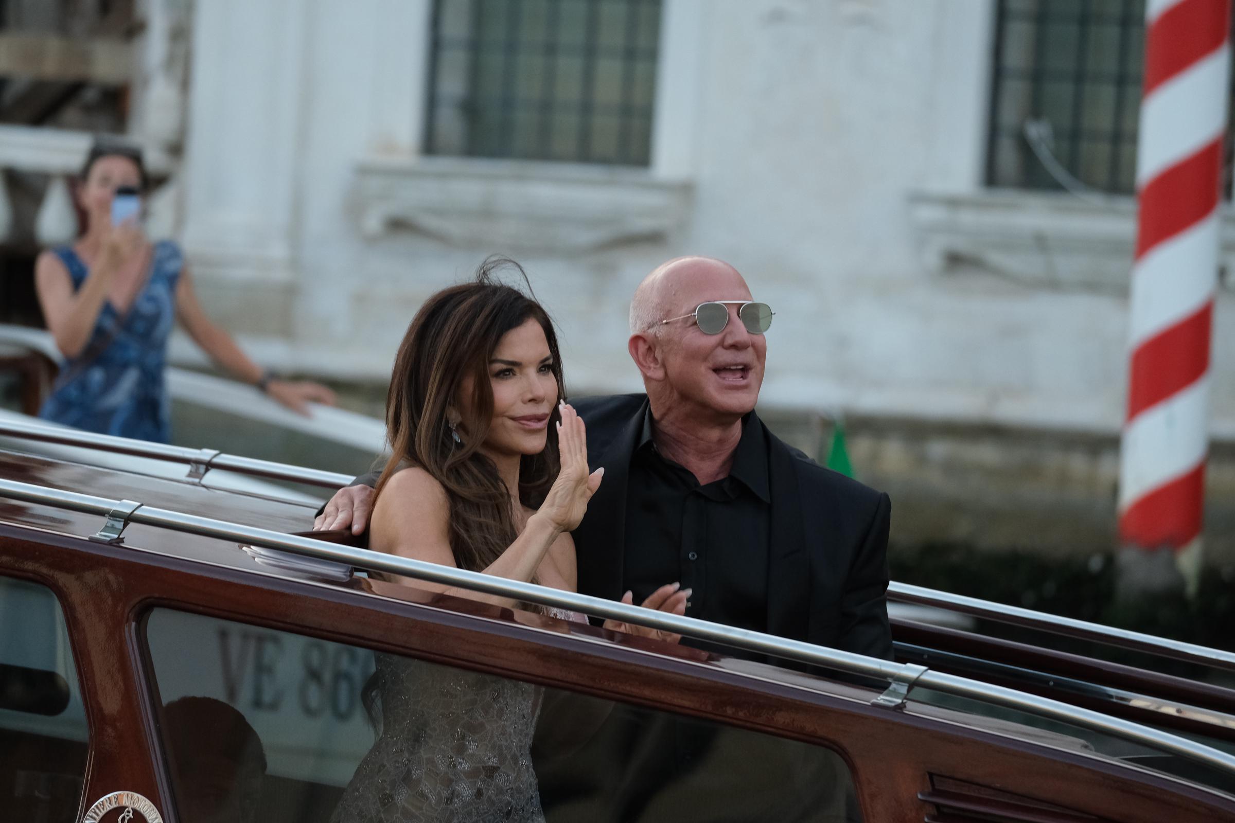 Lauren Sánchez hebt ihre Hand zu einem sanften Winken, während Jeff Bezos neben ihr lächelt. Beide stehen auf dem Boot, während eine Frau im Hintergrund den Moment mit ihrem Handy neben einem rot-weiß gestreiften Pfosten festhält.