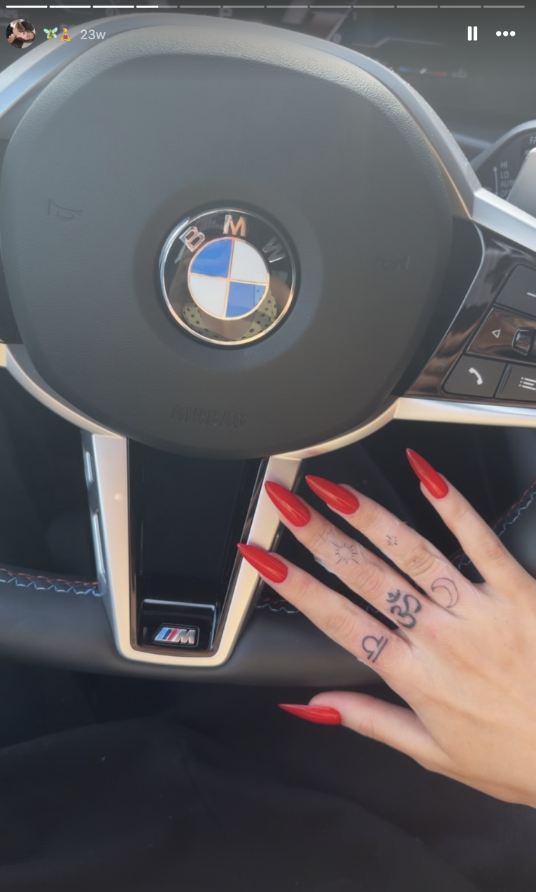 Harper Lockwood präsentiert ihre Sammlung zarter Fingertattoos – darunter ein heiliges Om-Symbol, himmlische Motive und das Sternzeichen Waage –, während sie ihre Hand mit spitzen roten Stiletto-Nägeln auf einem BMW-Lenkrad abstützt. | Quelle: Instagram/harper.lockwoodd