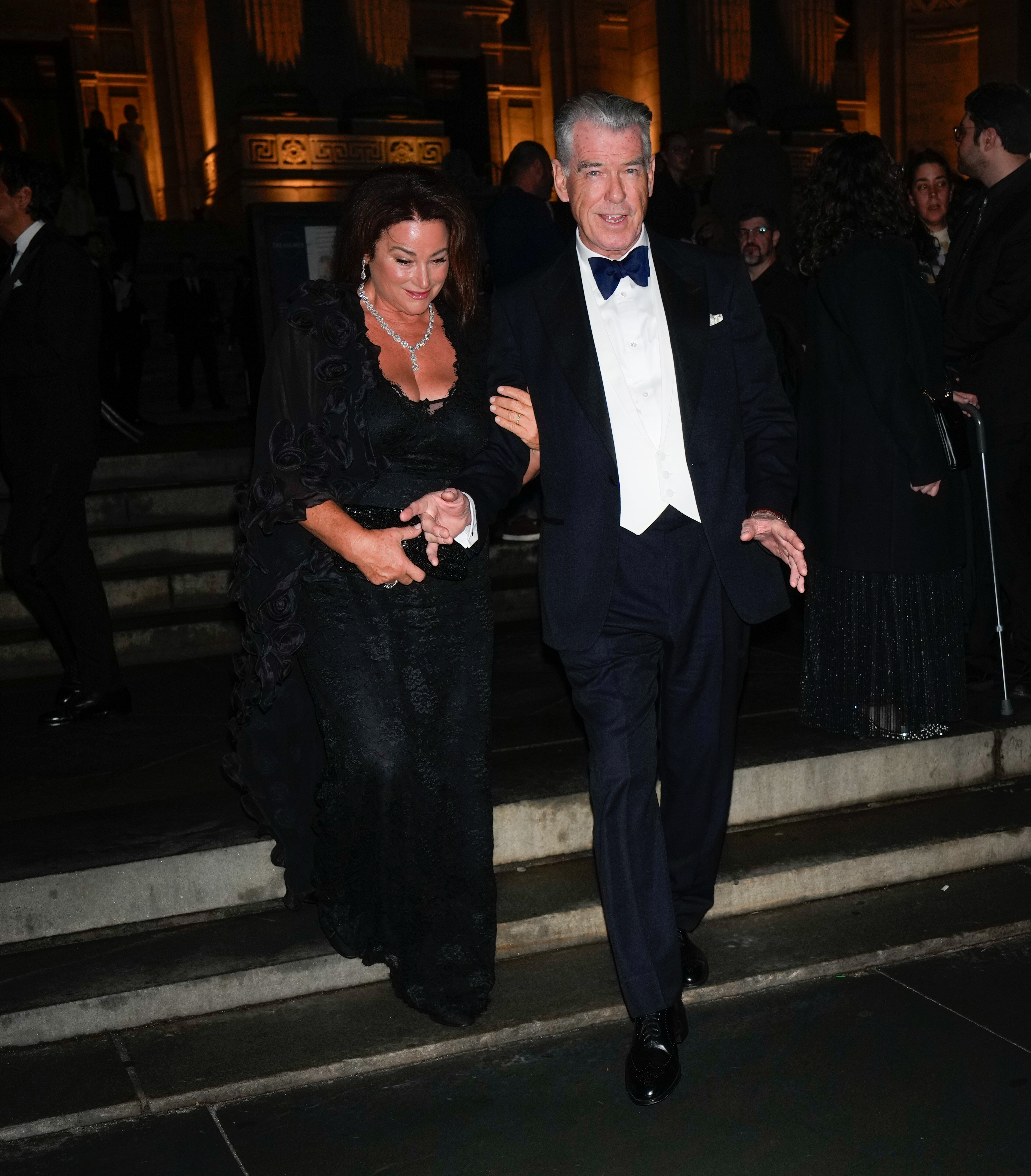 Pierce Brosnan hält sanft Keely Shaye Smiths Hand, während die beiden gemeinsam die Treppe hinuntergehen. Keely trägt ein elegantes schwarzes Spitzenkleid und funkelnden Schmuck. | Quelle: Getty Images.