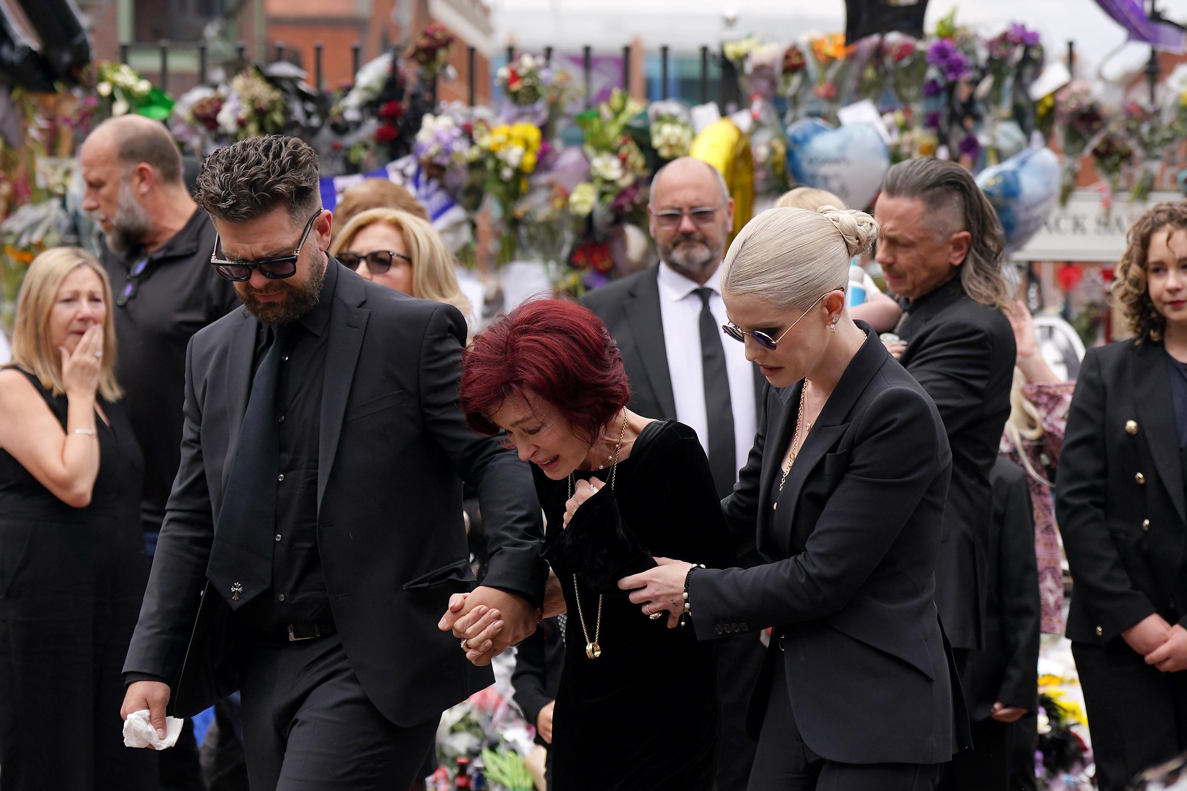 Jack, Sharon und Kelly Osbourne gehen langsam Hand in Hand auf die Botschaften und Blumengestecke zu, die am 30. Juli 2025 an der Black-Sabbath-Bank und der Black-Sabbath-Brücke in der Broad Street in Birmingham niedergelegt wurden. | Quelle: Getty Images