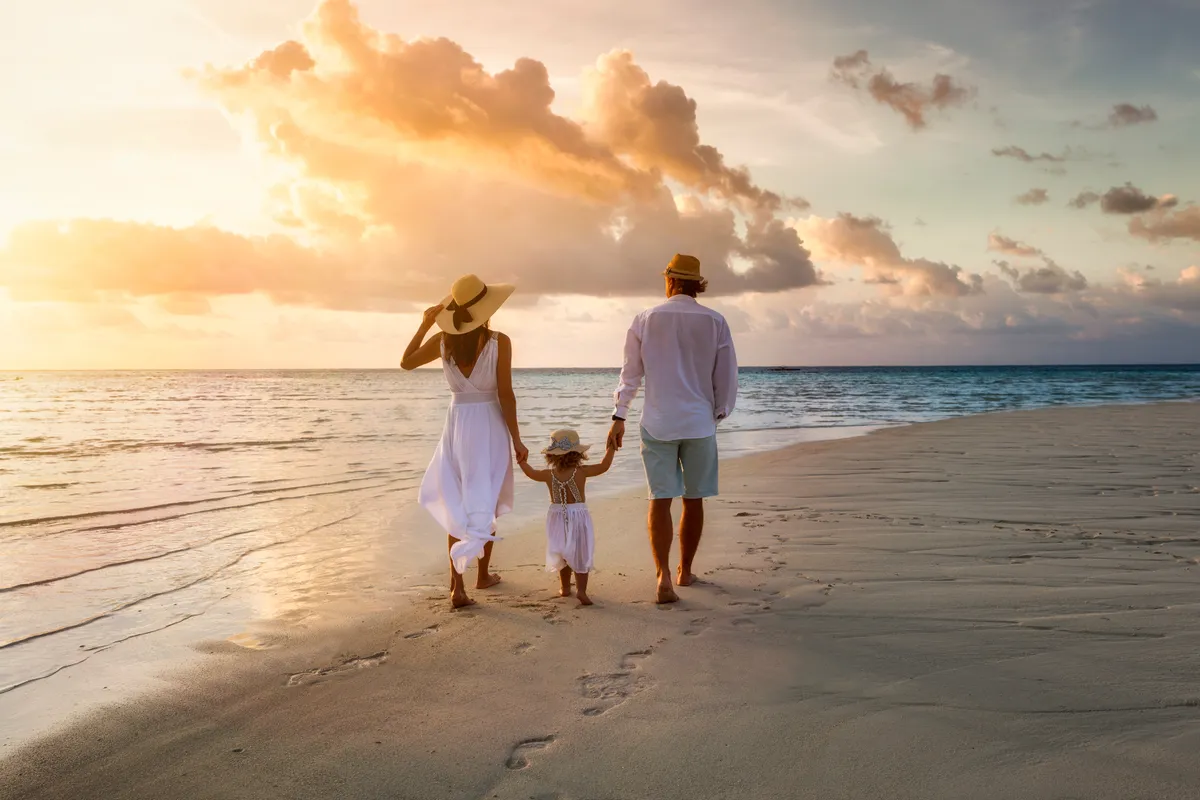 Eine elegante Familie in weißer Sommerkleidung spaziert Hand in Hand bei Sonnenuntergang an einem paradiesischen Strand entlang | Quelle: Shutterstock