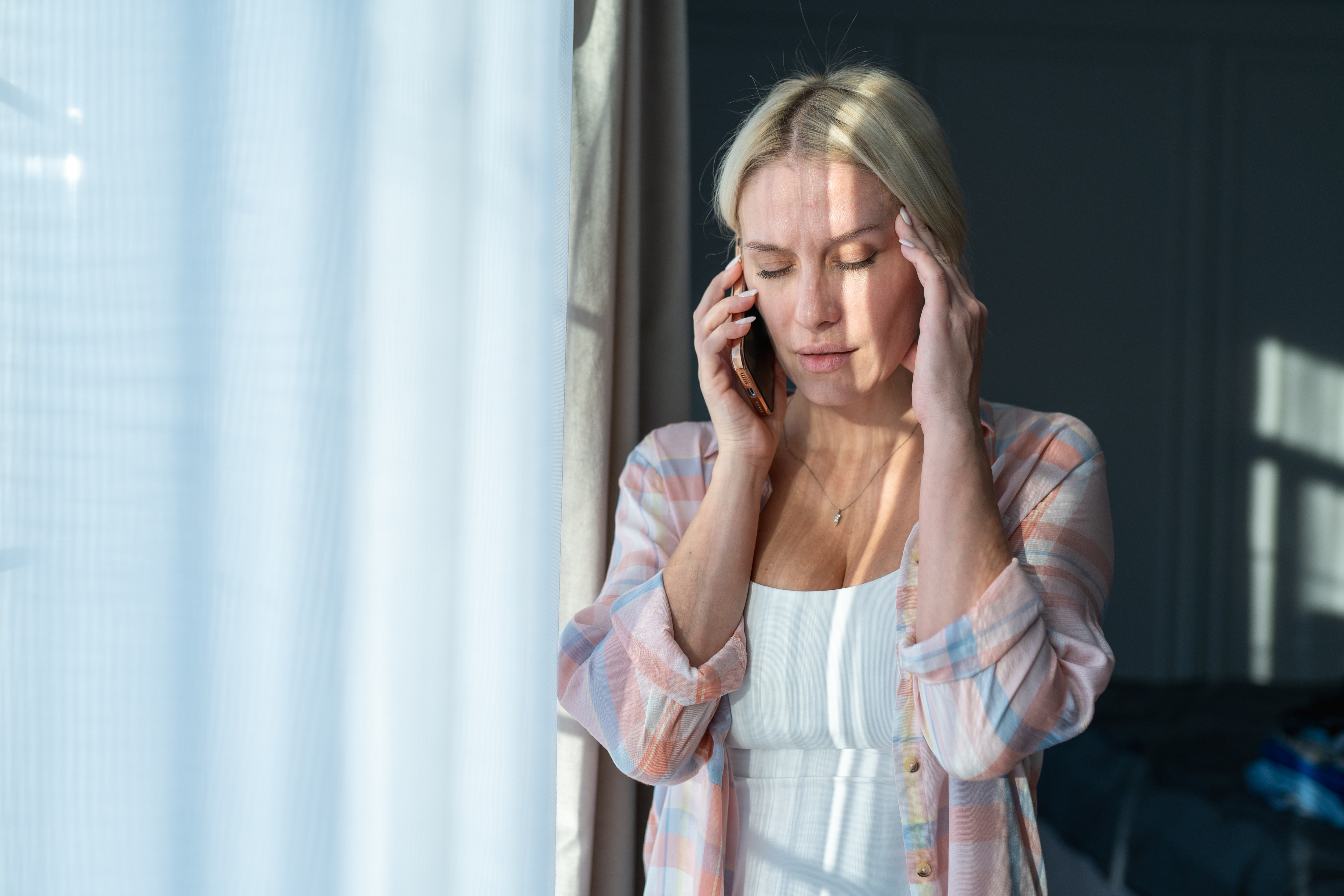 Kopfschmerzen und Verzweiflung. Blonde Frau telefoniert, während sie ihren Kopf am hellen Fenster berührt | Quelle: Getty Images