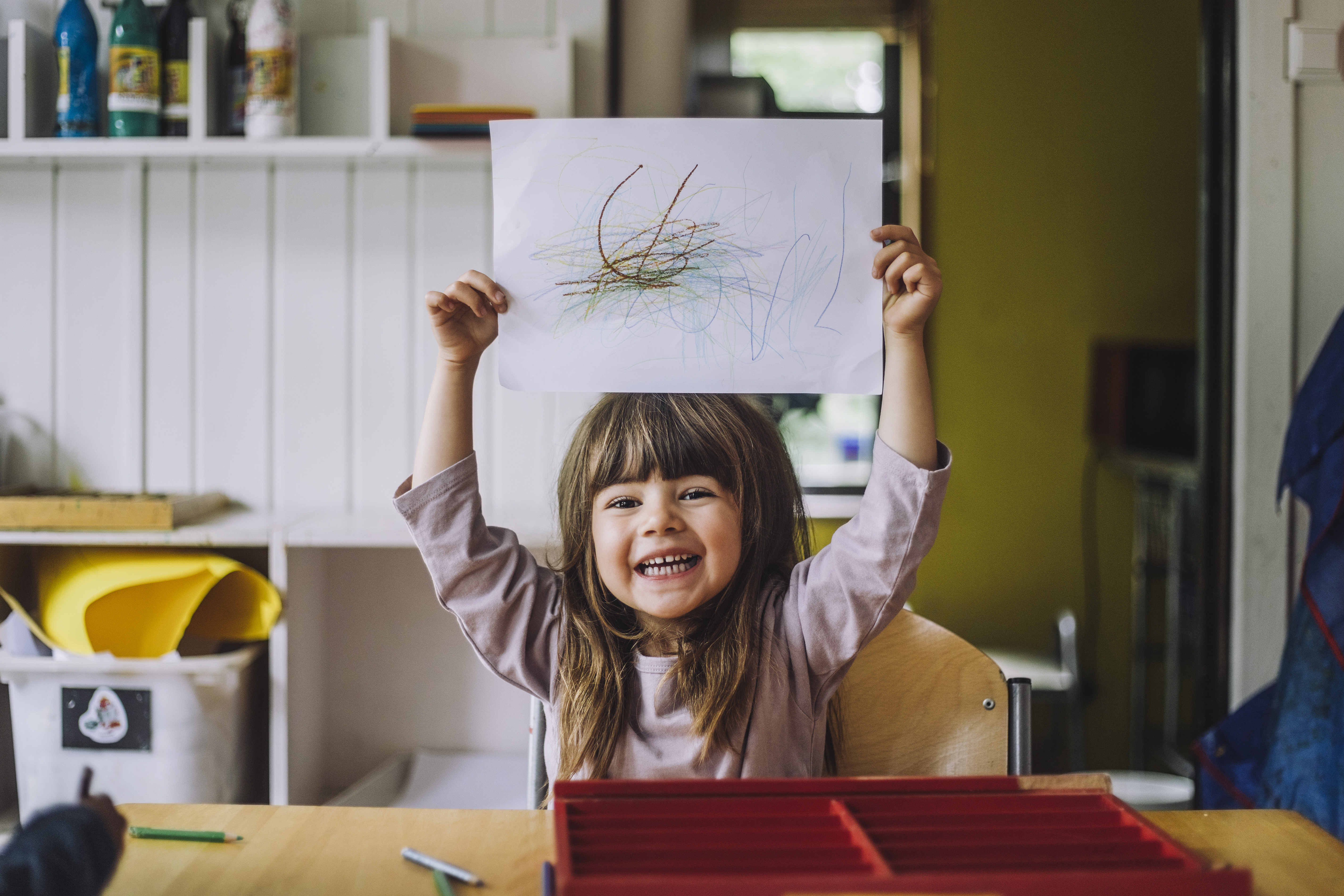 Fröhliches kleines Mädchen, das im Kindergarten mit Buntstiften auf Papier kritzelt | Quelle: Getty Images