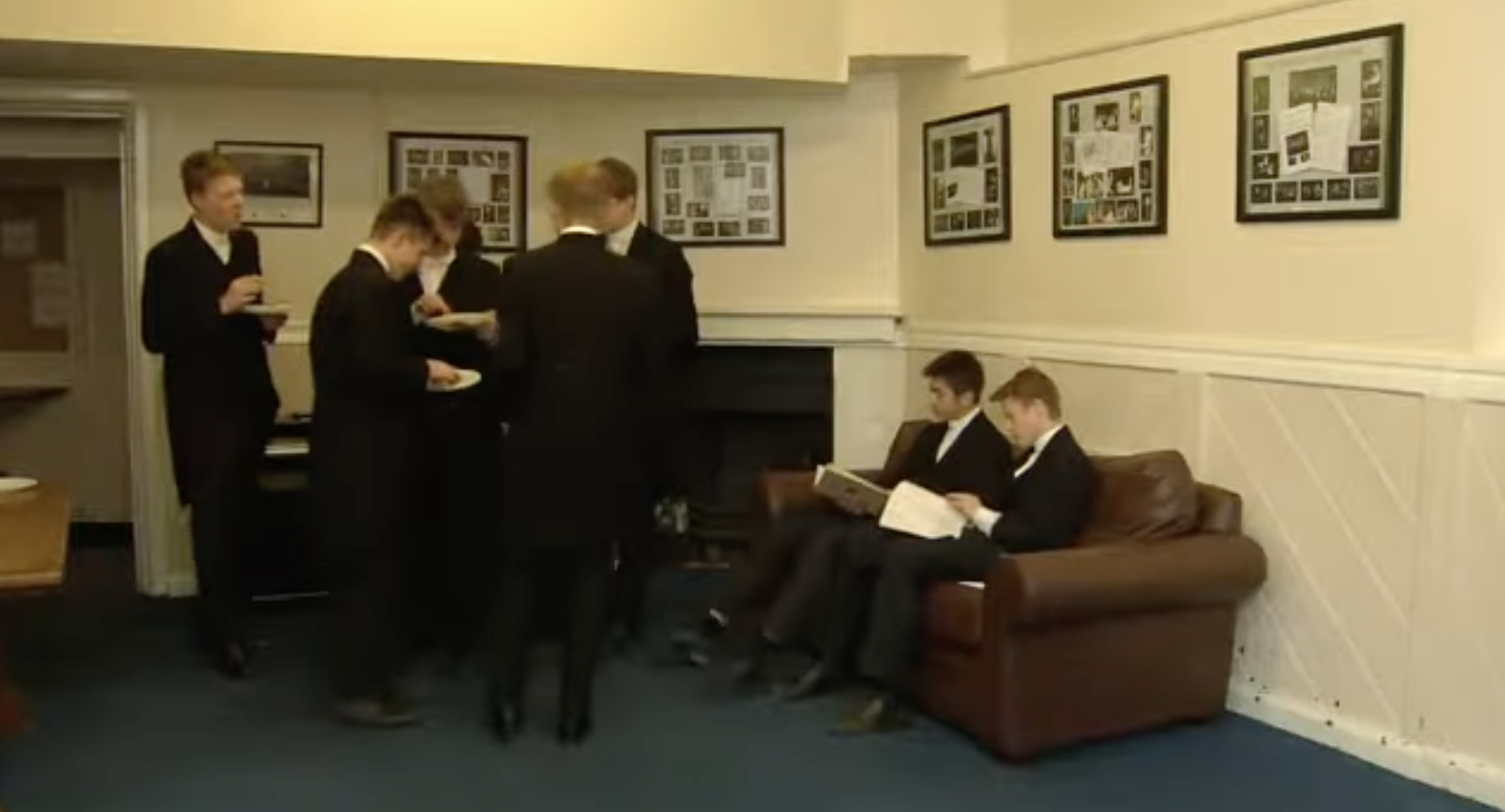 Die Schüler versammeln sich in einem Gemeinschaftsraum, der mit einem braunen Sofa, einem Kamin und gerahmten Bildern ausgestattet ist. Der Gemeinschaftsraum gibt einen Einblick in das Leben jenseits der einzelnen Schlafzimmer in Eton. | Quelle: YouTube/Paradoxyy