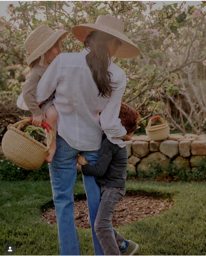 Meghan Markle spaziert durch einen blühenden Obstgarten, ein Kind auf dem Arm, während sich ein anderes an ihre Seite klammert – ein spontaner Moment aus dem Familienleben im Freien. | Quelle: Instagram/aseverofficial und meghan