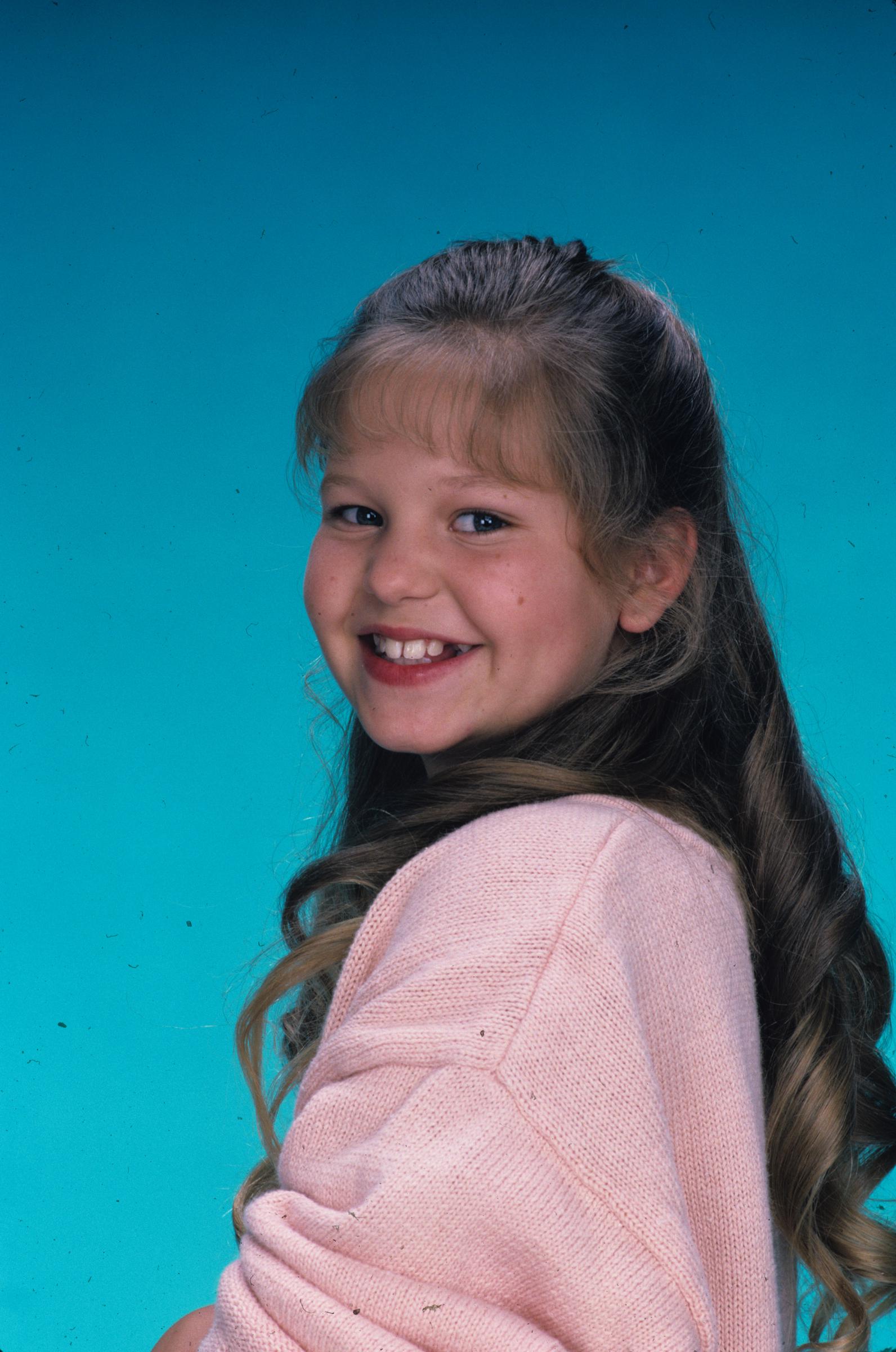 Die junge Candace Cameron strahlt während eines Werbe-Fotoshootings für die Pilotfolge „Our Very First Show“ der erfolgreichen ABC-Sitcom „Full House“ am 22. September 1987, in der sie die älteste Tochter D.J. Tanner spielte. Vor einem leuchtend türkisfarbenen Hintergrund posiert die Kinderdarstellerin in einem pastellrosa Strickpullover, ihr langes braunes Haar ist zu sanften Locken gestylt und wird von einem klassischen geraden Pony abgerundet. | Quelle: Getty Images