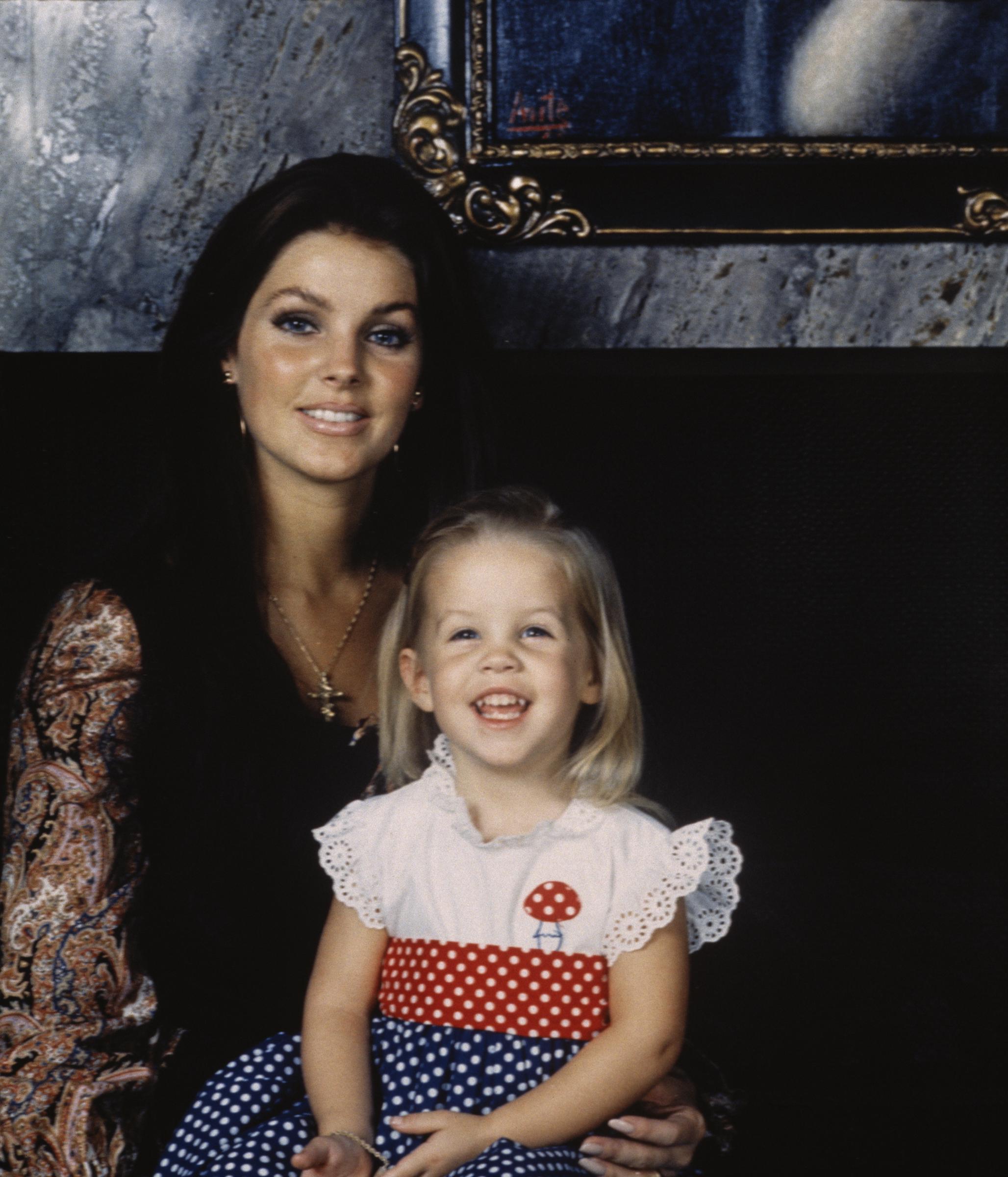 Priscilla Presley posiert mit ihrer kleinen Tochter, Lisa-Marie Presley, während der Thanksgiving-Woche im November 1970 in ihrem Haus in der Hillcrest Road in Beverly Hills, Kalifornien. Sie trägt ein dunkles Oberteil mit bunten Paisley-Ärmeln und eine goldene Kreuzkette, während Lisa-Marie in einem weißen, mit Lochstickerei verzierten Polka-Dot-Kleid mit rotem Pilzmotiv lächelt. | Quelle: Getty Images