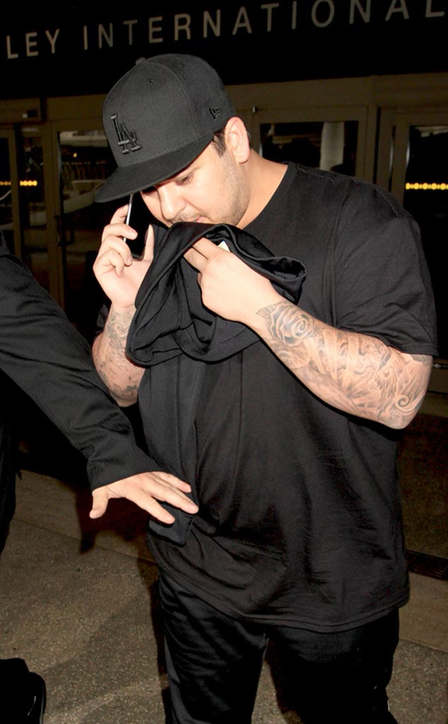 Rob Kardashian wird vor einem Flughafen fotografiert, wie er sich ein schwarzes T-Shirt vor einen Teil seines Gesichts hält, während er telefoniert. Das Bild erinnert an sein schwieriges Verhältnis zur Aufmerksamkeit der Paparazzi, das – wie er später verriet – zu seiner Entscheidung beigetragen hatte, Kim Kardashians Hochzeit im Jahr 2014 zu verpassen. | Quelle: Getty Images.