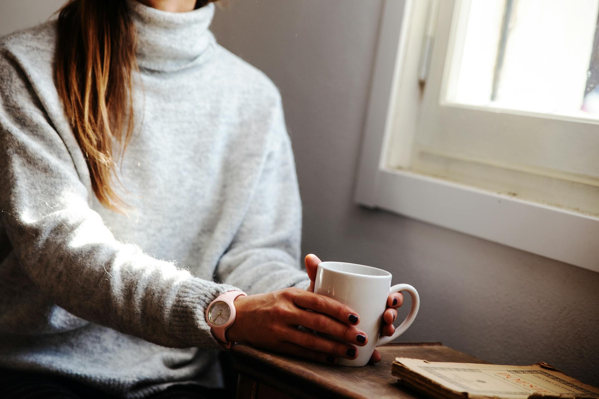 Eine Frau hält einen Kaffeebecher und sitzt am Fenster | Quelle: Pexels