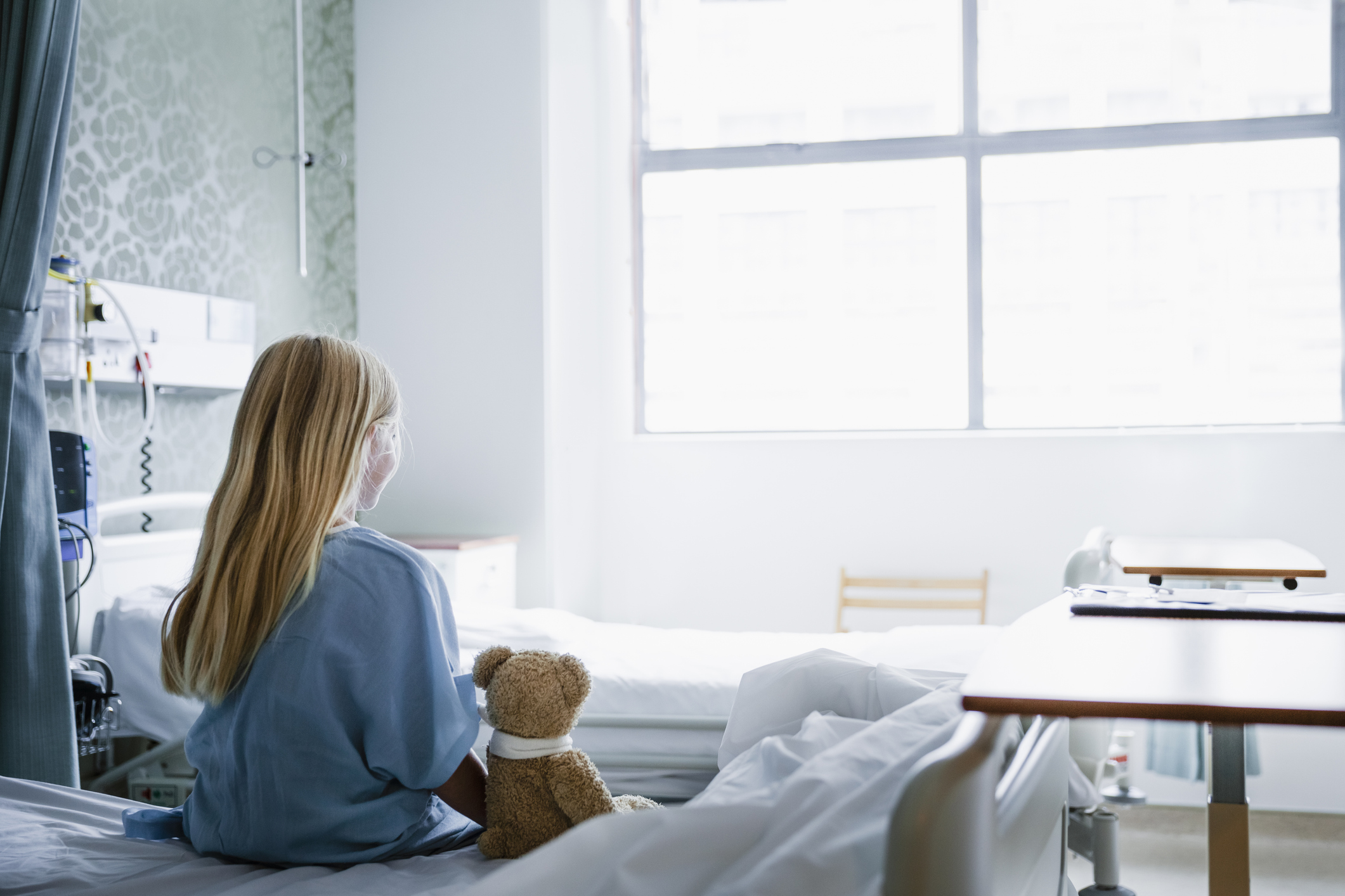Kleines Mädchen sitzt auf einem Krankenhausbett, neben ihr liegt ihr Teddybär, und sie schaut zum Fenster hinaus. | Quelle: Getty Images