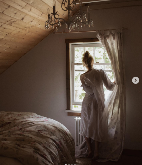 Von hinten gesehen, in einem ätherisch weißen Nachthemd und mit zu einem Dutt hochgestecktem Haar, zieht Pamela Anderson einen gemusterten Vorhang beiseite, um aus einem sonnendurchfluteten Dachfenster in Arcady hinauszuschauen. In sanftes Morgenlicht getaucht, unter einem Kristallleuchter und einer schrägen Holzdecke, spiegelt das gemütliche Schlafzimmer im Dachgeschoss perfekt ihre Bildunterschrift wider: „Der Herbst ist eine wunderschöne Jahreszeit … erholsam, friedlich … geborgen, flauschige Socken … sich einnisten, Winterschlaf halten … 🤍“. | Quelle: Instagram/pamelaanderson