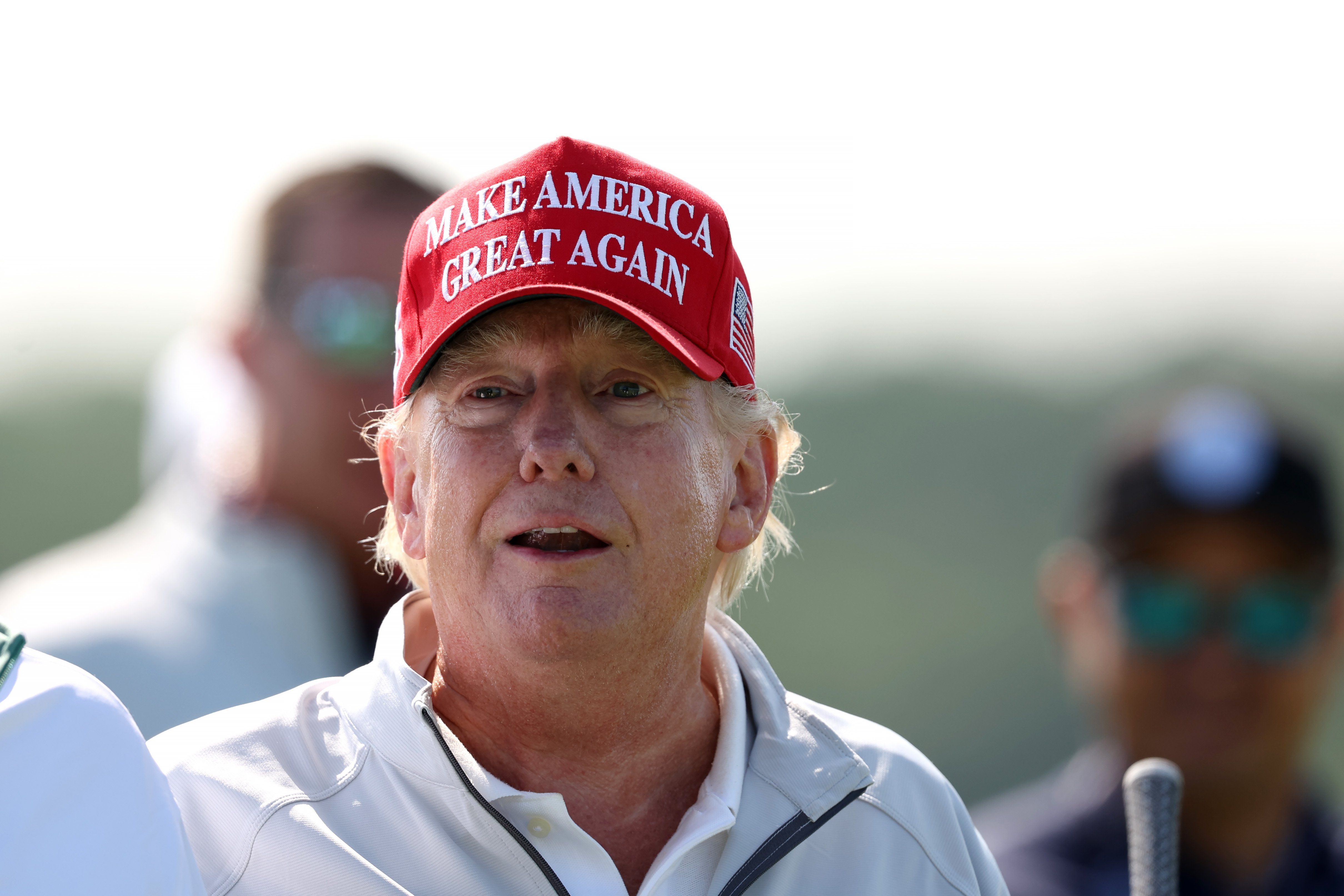 Beim LIV Golf Invitational in Sterling, Virginia, steht Donald Trump am 25. Mai im Sonnenlicht und trägt eine rote „Make America Great Again“-Kappe und eine weiße Jacke mit Kragen. Seine Wangen und seine Nase sind stark gerötet, während sein Hals viel heller aussieht.
