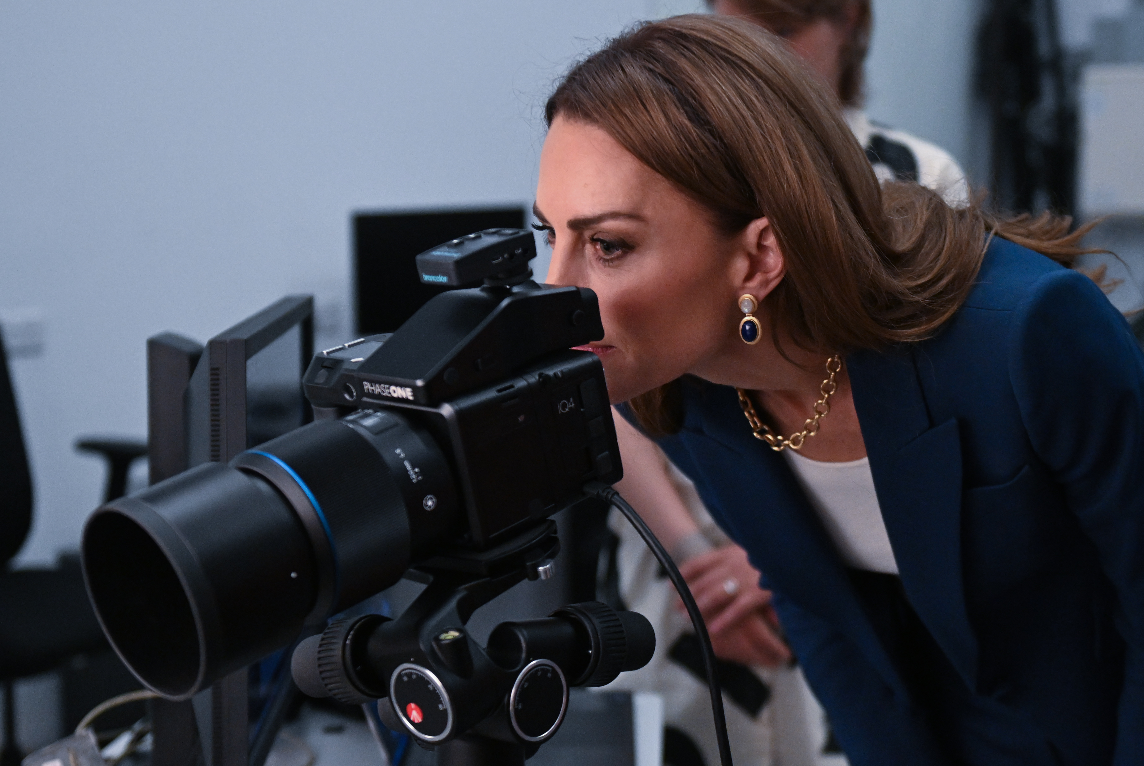 Konzentriert und neugierig beugt sich Prinzessin Catherine vor, um eine Kameraeinstellung zu beobachten. Ihr blauer Blazer und die passenden Ohrringe heben sich von dem minimalistischen Raum ab, während ihr glattes Haar und ihre Accessoires einen ruhigen, modernen Look vervollständigen.