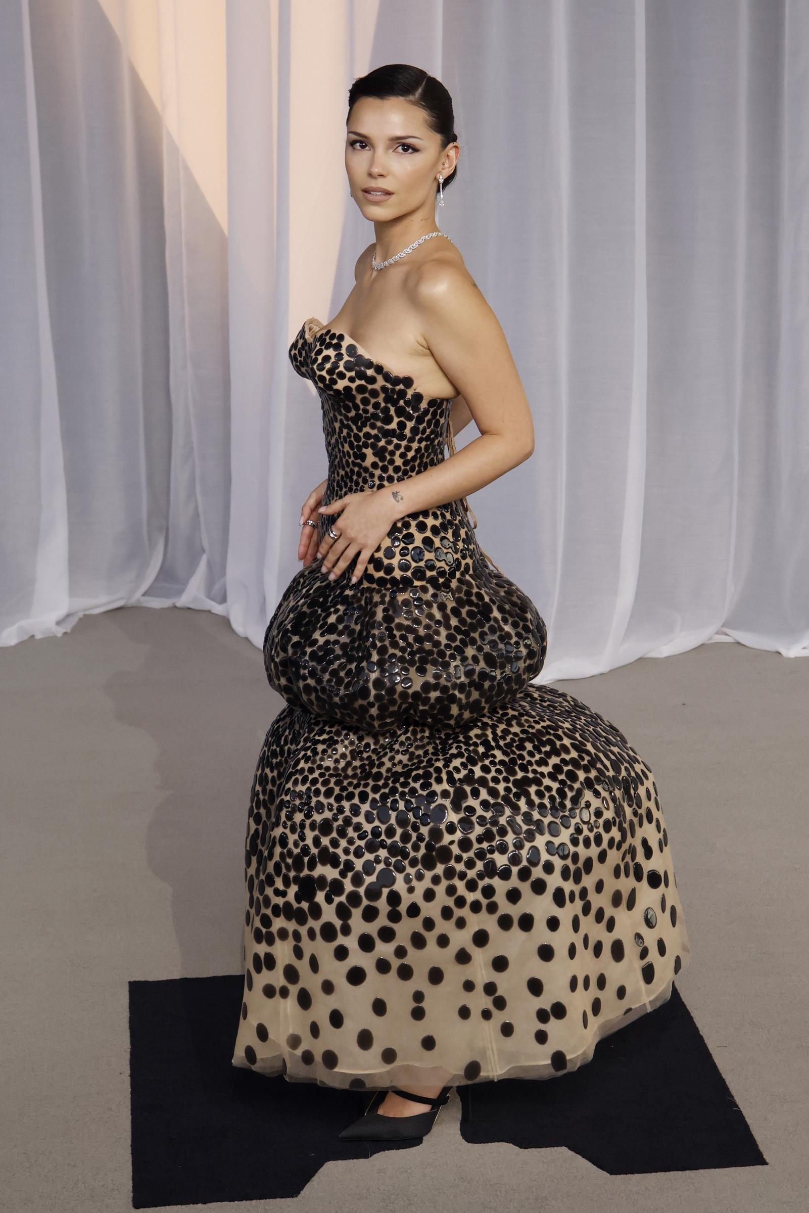 Inde Navarrette sorgt für ein auffälliges, avantgardistisches Drama in einem figurbetonten, trägerlosen Kleid, das mit glänzenden, übergroßen Polka-Dots auf einem neutralen Hintergrund verziert ist. Die voluminöse Silhouette weitet sich kühn an den Hüften und am gestuften Bubble-Saum aus, was sie mit einer glatten, eleganten Hochsteckfrisur, einem funkelnden Diamant-Choker und markanten schwarzen Pumps mit spitzer Zehenpartie ausgleicht.