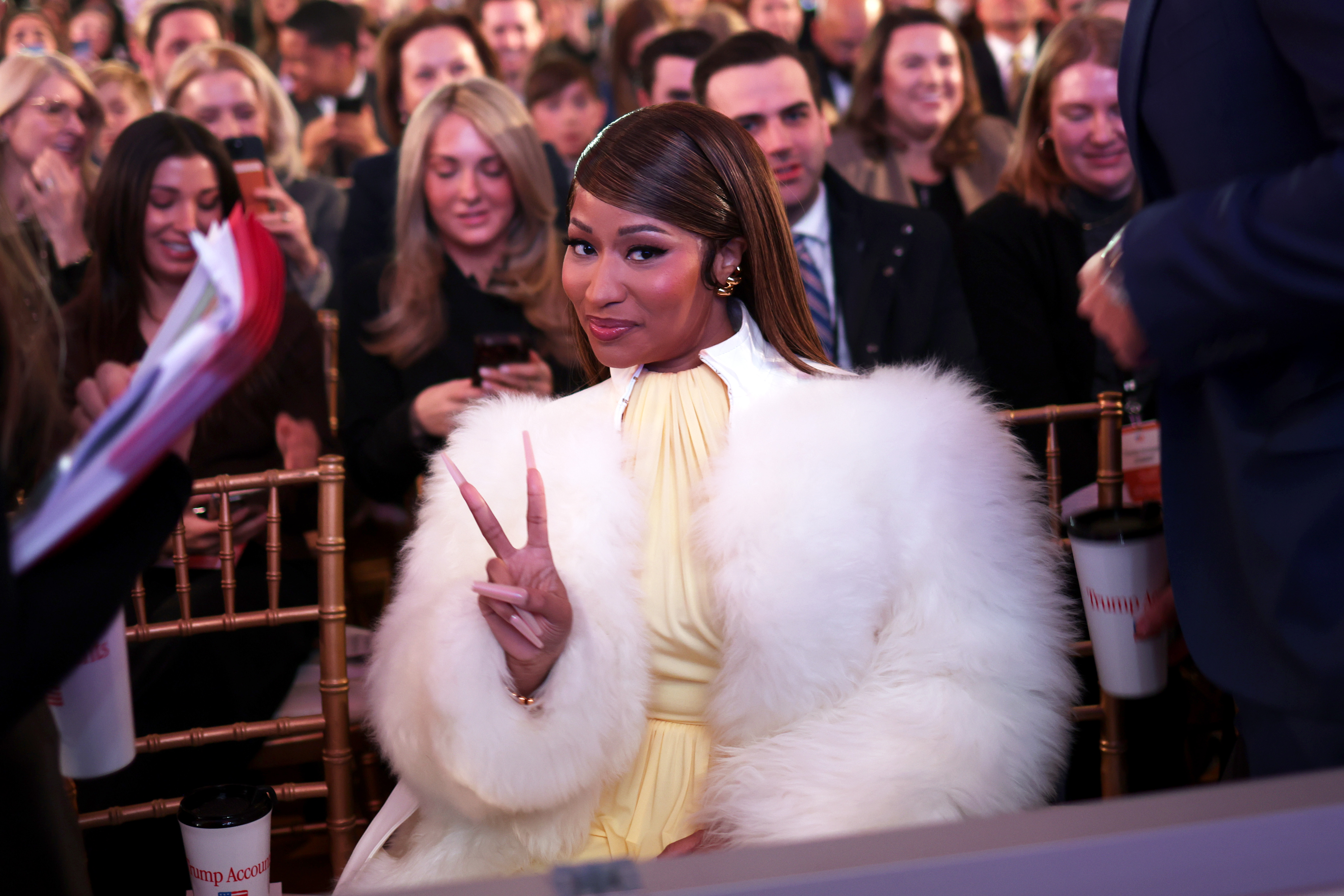 Nicki Minaj zeigt das Peace-Zeichen, während sie bei einer Veranstaltung von „Trump Accounts“ sitzt – sie trägt einen voluminösen weißen Pelzmantel über einem hellgelben Outfit. Dieser Auftritt war ein weiteres Beispiel dafür, wie sie glamouröse Mode mit eher formellen öffentlichen Anlässen kombiniert. | Quelle: Getty Images