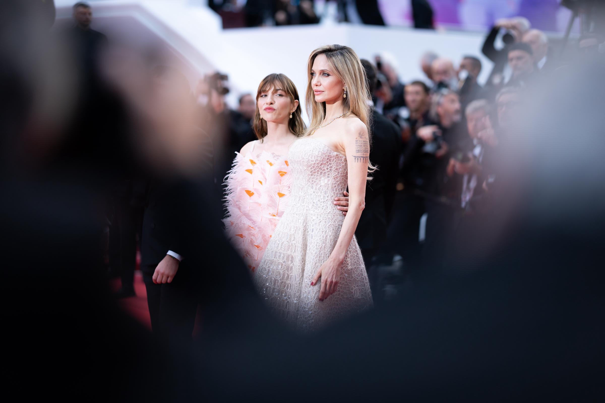 Eingerahmt von verschwommenen Silhouetten im Vordergrund, teilen Angelina Jolie und Marie Colomb einen auffälligen Moment auf dem roten Teppich. Jolie steht in ihrer paillettenbesetzten Cucinelli-Robe aufrecht da, während Colomb in einem mit Federn besetzten, von Blumen inspirierten Minikleid die Blicke auf sich zieht - beide Frauen ziehen die Blicke auf sich.