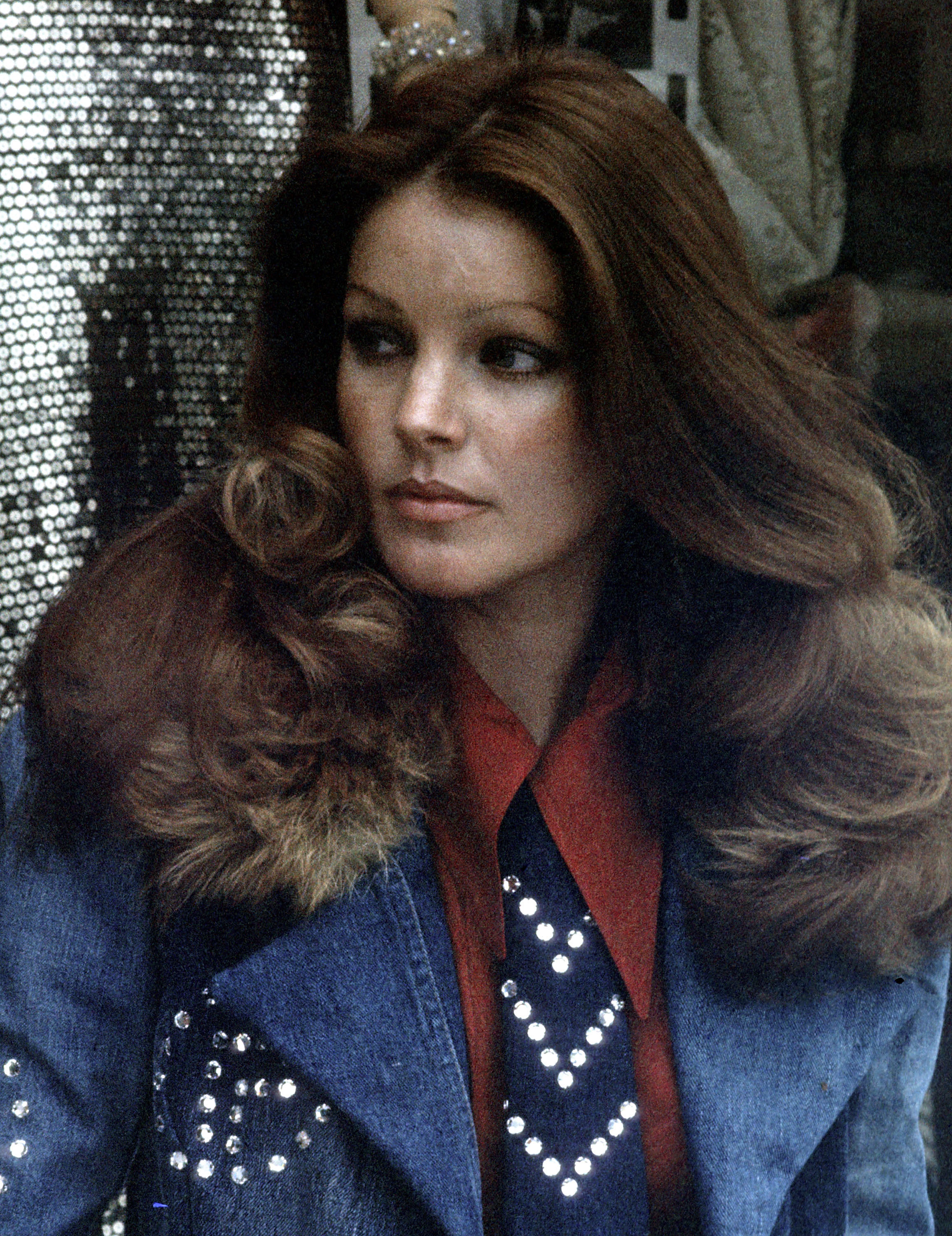 Priscilla Presley posiert während eines Fotoshootings in ihrem Bekleidungsgeschäft „Bis & Beau Boutique“ in Beverly Hills, Kalifornien, am 19. März 1974. Sie trägt eine Jeansjacke mit Pelzkragen und Strassverzierungen über einem rostroten Hemd mit Kragen und einer Krawatte mit Nieten, während ihr rotbraunes Haar zu voluminösen, gestuften Wellen gestylt ist. | Quelle: Getty Images