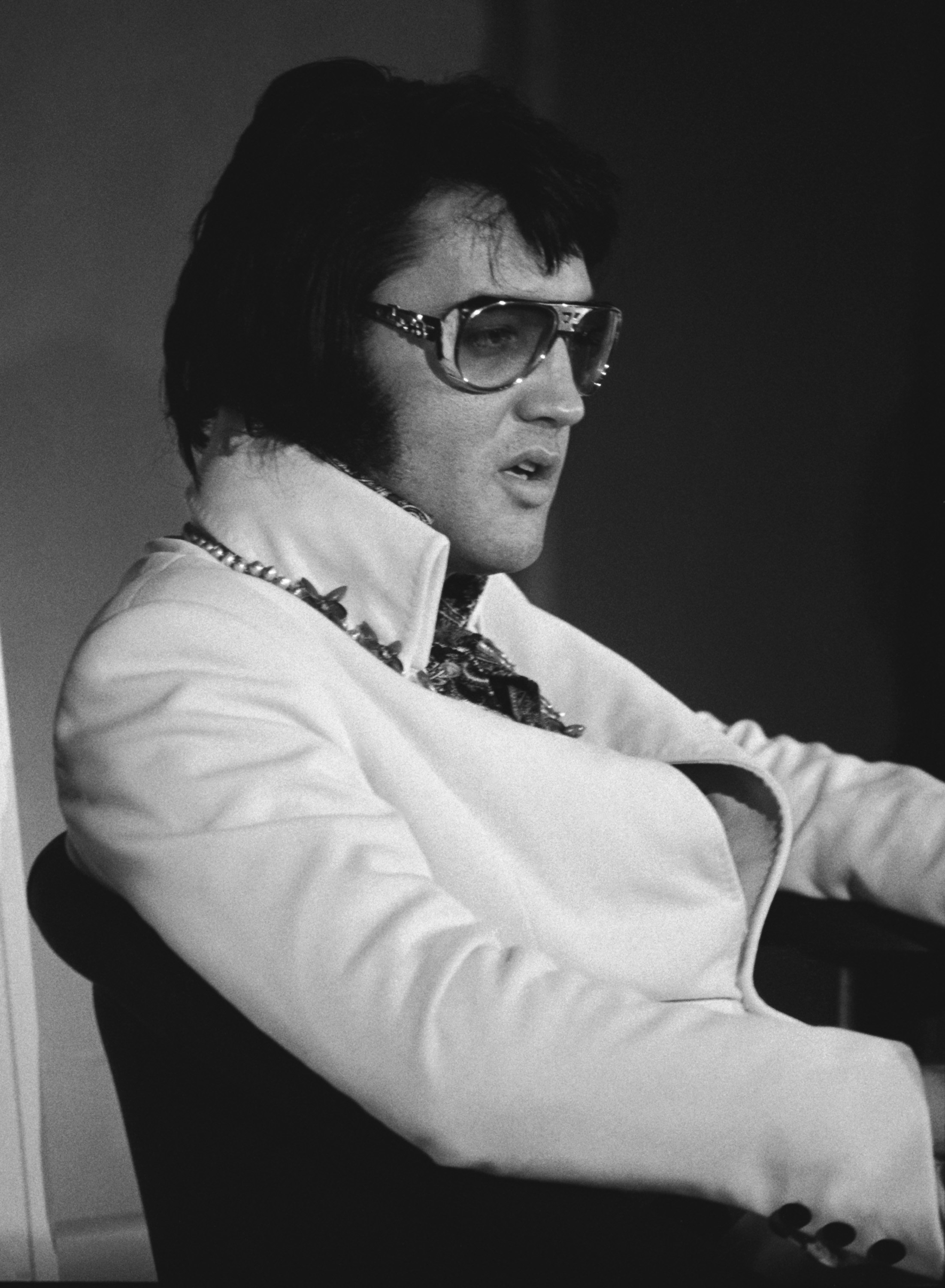 Elvis Presley nimmt am 4. September 1972 im Las Vegas Hilton in Las Vegas, Nevada, an einem Werbeinterview für sein bevorstehendes Fernsehkonzert „Elvis: Aloha from Hawaii“ teil. Er trägt eine offene, hellfarbige Jacke mit hohem Kragen über einem aufwendig gemusterten Ascot und einer Kettenhalskette, dazu seine typische übergroße, getönte Pilotenbrille und Koteletten.
