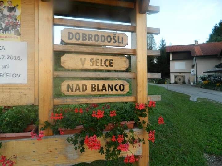 Ein rustikales hölzernes Ortsschild am Straßenrand, geschmückt mit leuchtend roten Geranien, begrüßt Besucher mit dem eingeschnitzten Gruß „DOBRODOŠLI V SELCE NAD BLANCO" in der Ortschaft Selce nad Blanco in der Gemeinde Sevnica, Slowenien. Das friedliche Ortseingangsschild rahmt einen Blick auf grüne Hügel, eine örtliche Straße und Wohnhäuser in der ländlichen Gemeinde. | Quelle: Facebook/Turistična kmetija Rostohar