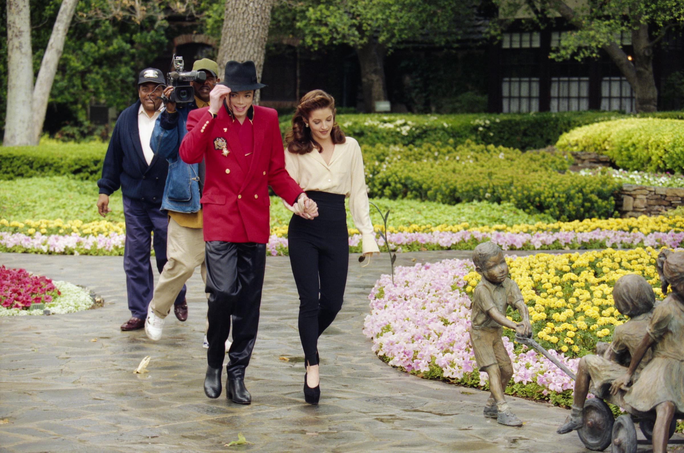 Lisa Marie Presley hält Händchen mit ihrem Ehemann Michael Jackson, während die beiden durch die blumengeschmückten Anlagen der Neverland Ranch schlendern, um sich auf den Children’s World Summit am 18. April 1995. Sie trägt eine elfenbeinfarbene, langärmelige Seidenbluse, die sie in eine schwarze, hoch taillierte, schmal zulaufende Hose gesteckt hat, dazu High Heels, während Jackson seinen schwarzen Fedora zurechtzieht – er trägt einen leuchtend roten, zweireihigen Blazer im Militärstil und eine schwarze Hose. | Quelle: Getty Images