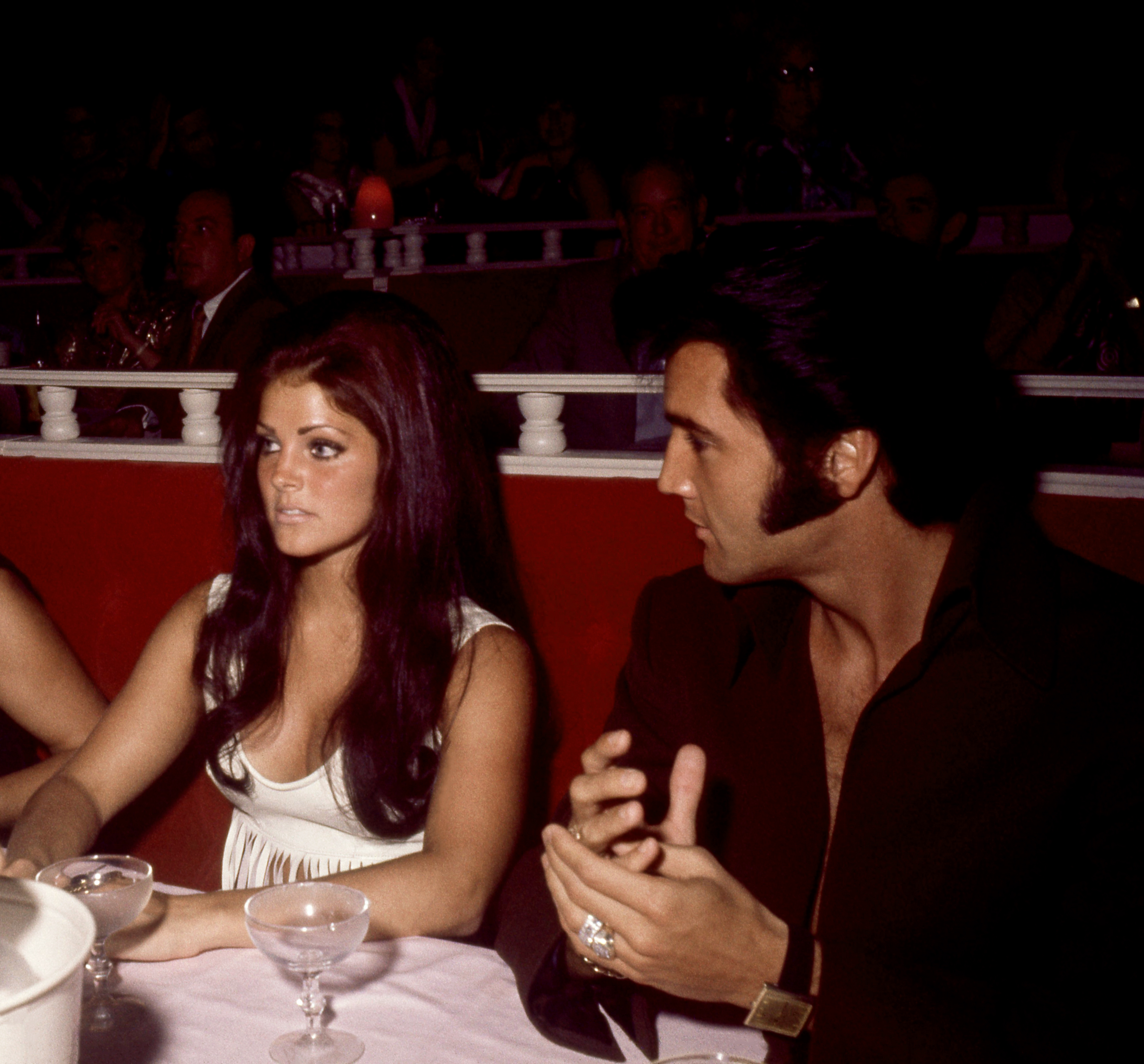 Priscilla Presley sitzt neben ihrem Ehemann Elvis Presley an einem Banketttisch vor seinem Eröffnungsauftritt im International Hotel in Las Vegas, Nevada, im August 1969. Sie trägt ein ärmelloses weißes Oberteil mit U-Ausschnitt und hat ihr langes dunkles Haar zu voluminösen Wellen gestylt, während Elvis ein dunkles, aufgeknöpftes Hemd mit Kragen trägt. | Quelle: Getty Images