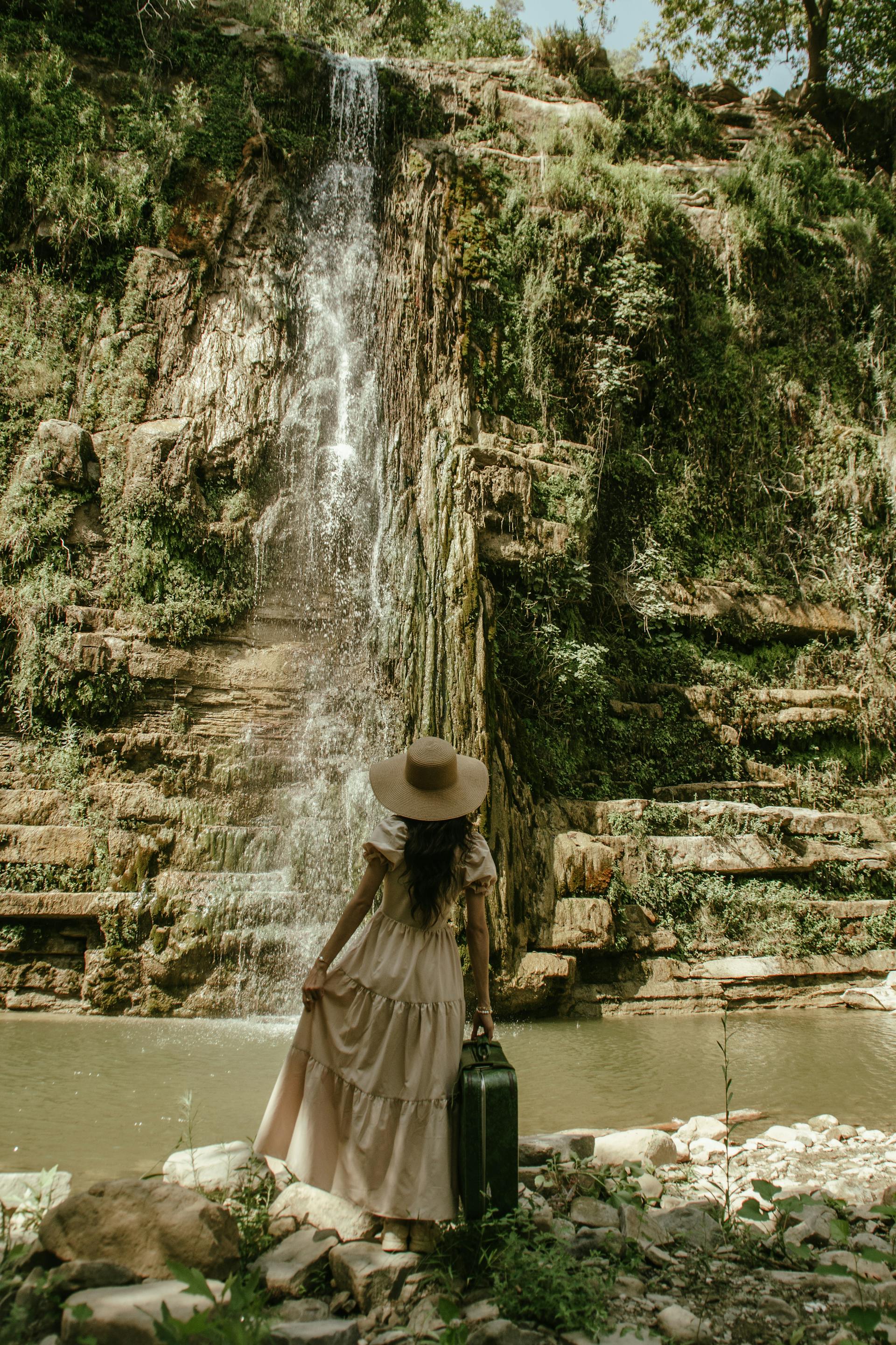 Eine Frau in einem beigen Kleid und einem Strohhut schaut auf einen Wasserfall | Quelle: Pexels