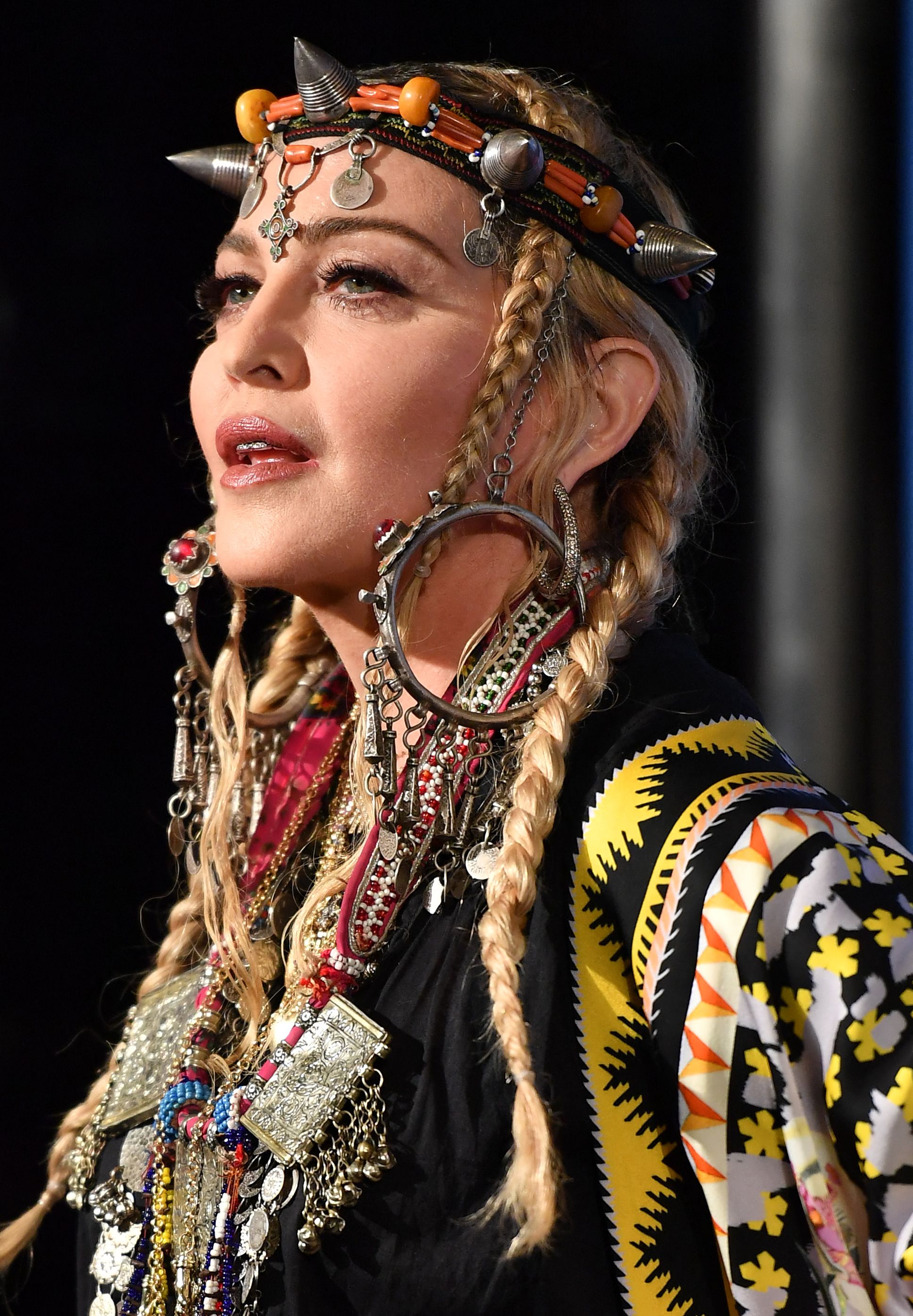 Madonna wird von der Seite fotografiert – mit langen blonden Zöpfen, einem verzierten Kopfschmuck, übergroßen Ohrringen und mehreren Lagen aufwendig gestalteter Schmuckstücke. | Quelle: Chat GPT