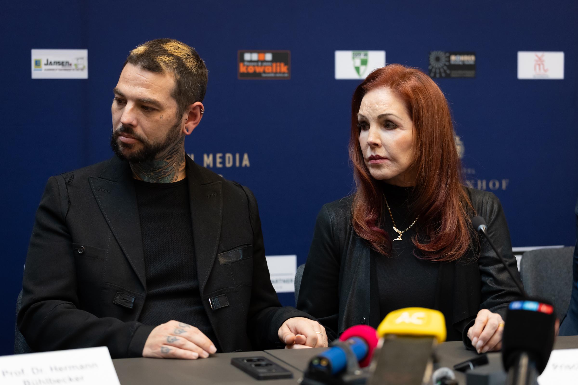 Priscilla Presley sitzt neben ihrem Sohn Navarone Garibaldi Garcia bei einer Presseveranstaltung in Baesweiler, Deutschland, am 20. Oktober 2026. Beide tragen aufeinander abgestimmte schwarze Kleidung, während sie hinter den Mikrofonen an einem Podiumstisch aufmerksam zuhören. Presley trägt eine goldene Halskette über einem schwarzen Oberteil und trägt ihr stufig geschnittenes rotbraunes Haar in sanften Wellen um die Schultern. | Quelle: Getty Images
