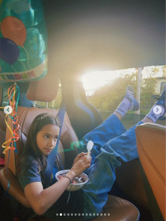Auf dem Foto sitzt Fiona allein auf dem Rücksitz eines Autos. Sie trägt ein dunkelblaues T-Shirt und blaue Jeans, dazu weiße Socken und Turnschuhe. Mit ausgestreckten Beinen auf dem Sitz hält sie einen Löffel über einen kleinen Behälter mit Essen auf ihrem Schoß, während Sonnenlicht durch das Fenster scheint und daneben bunte Luftballondekorationen hängen. | Quelle: Instagram/mabellowe_