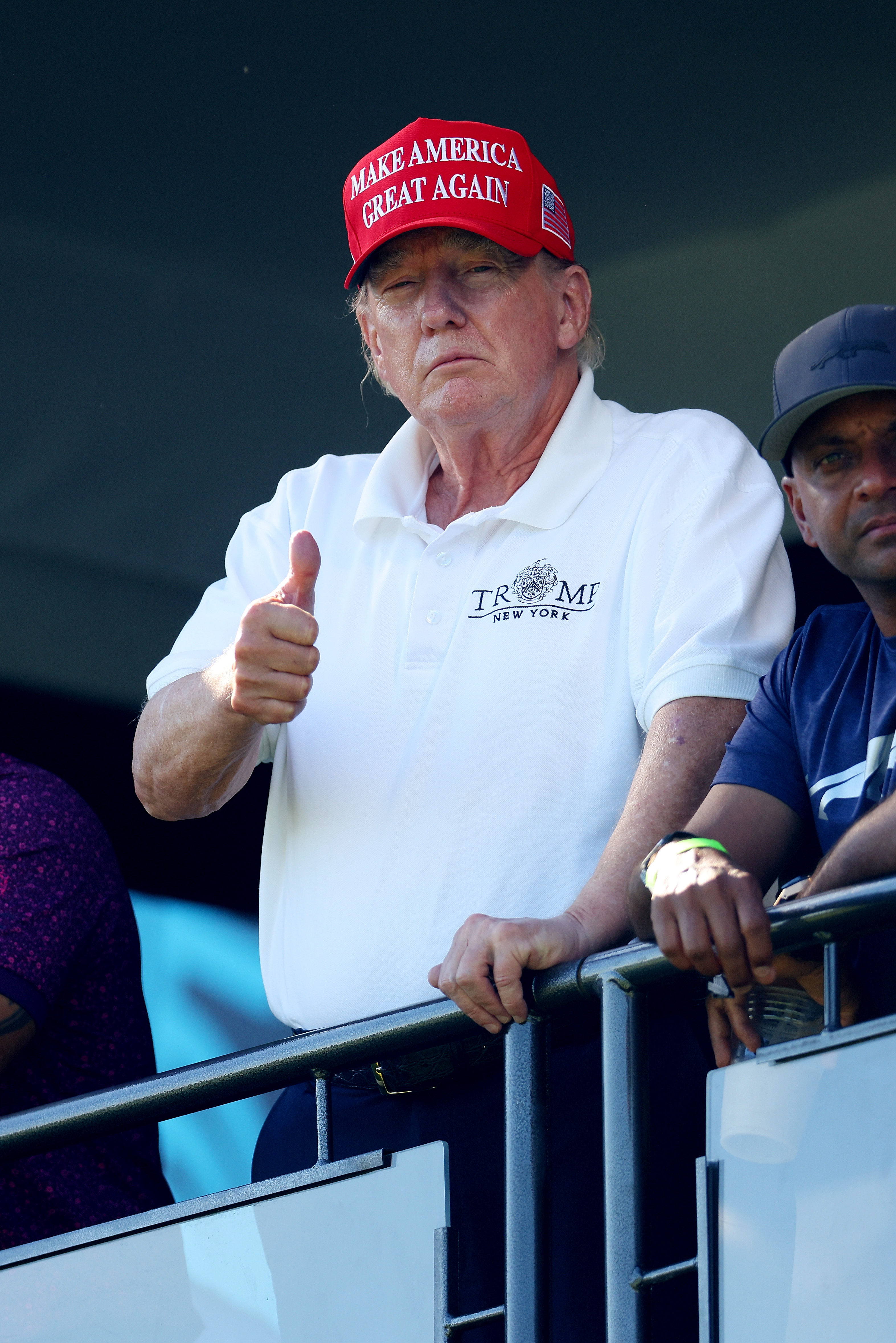 Am 13. August steht Donald Trump am Geländer des 18. Grüns und zeigt den Daumen nach oben. Die Rötung in seinem Gesicht bleibt sichtbar, mit helleren Tönen an den Rändern seines Kiefers und Halses.