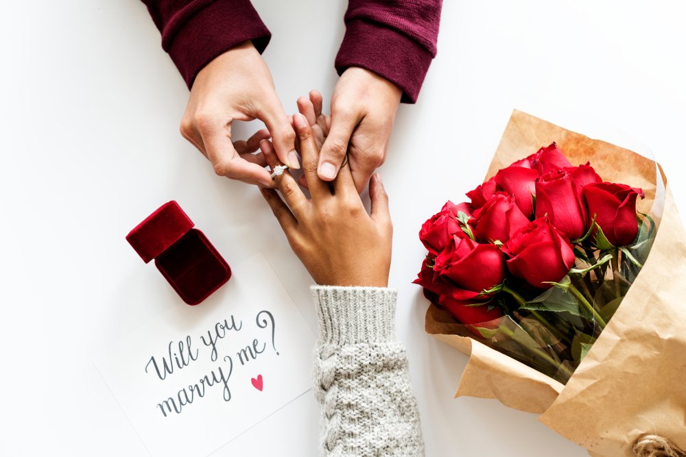 Mann steckt den Ring auf den Finger der Verlobten, neben einer Notiz auf der die Frage "Willst du mich heiraten", geschrieben wurde. | Quelle: Shutterstock  
