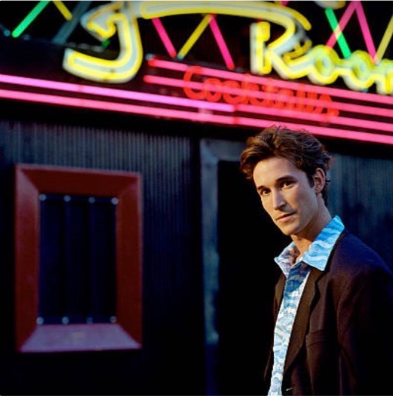 Auf einem alten Foto aus seinen frühen Hollywood-Jahren posiert der Schauspieler als junger Erwachsener vor einem neonbeleuchteten Veranstaltungsort – in einem gemusterten Hemd und einem dunklen Blazer. | Quelle: Instagram/therealnoahwyle
