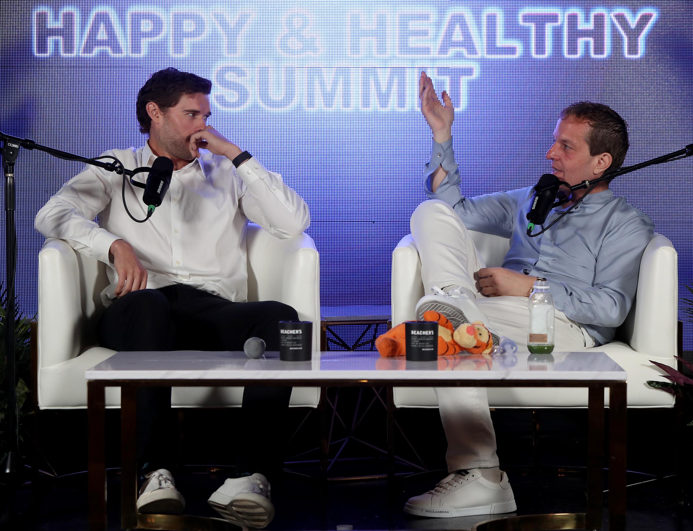 Christopher Schwarzenegger hört nachdenklich zu, als Jeff Beacher während ihrer Podiumsdiskussion einen Standpunkt vertritt. Die beiden sitzen sich auf der Bühne gegenüber und diskutieren intensiv über persönliches Wachstum und Gesundheit.