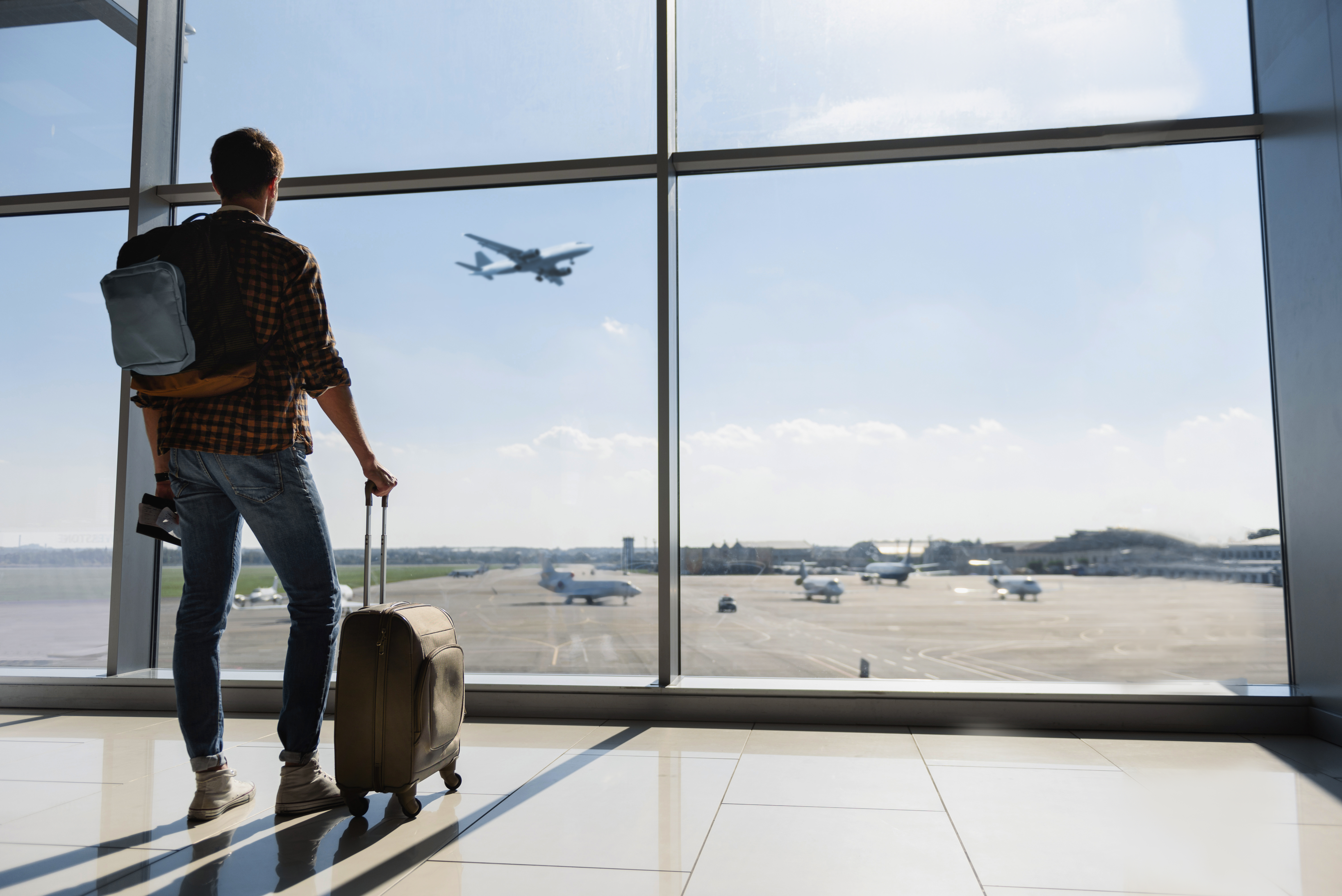 Ein junger Mann steht mit seinem Gepäck an einem Flughafenfenster und beobachtet ein Flugzeug vor dem Abflug | Quelle: Shutterstock